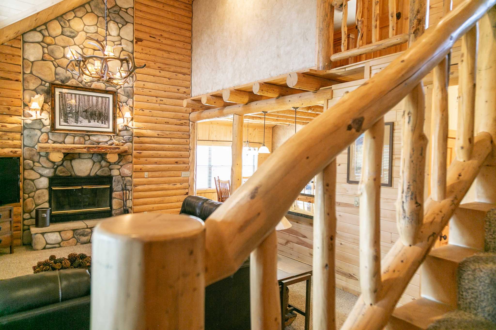 Interior of a log cabin living room with a wooden staircase in the foreground, a stone fireplace with a picture above, and large windows with natural light.