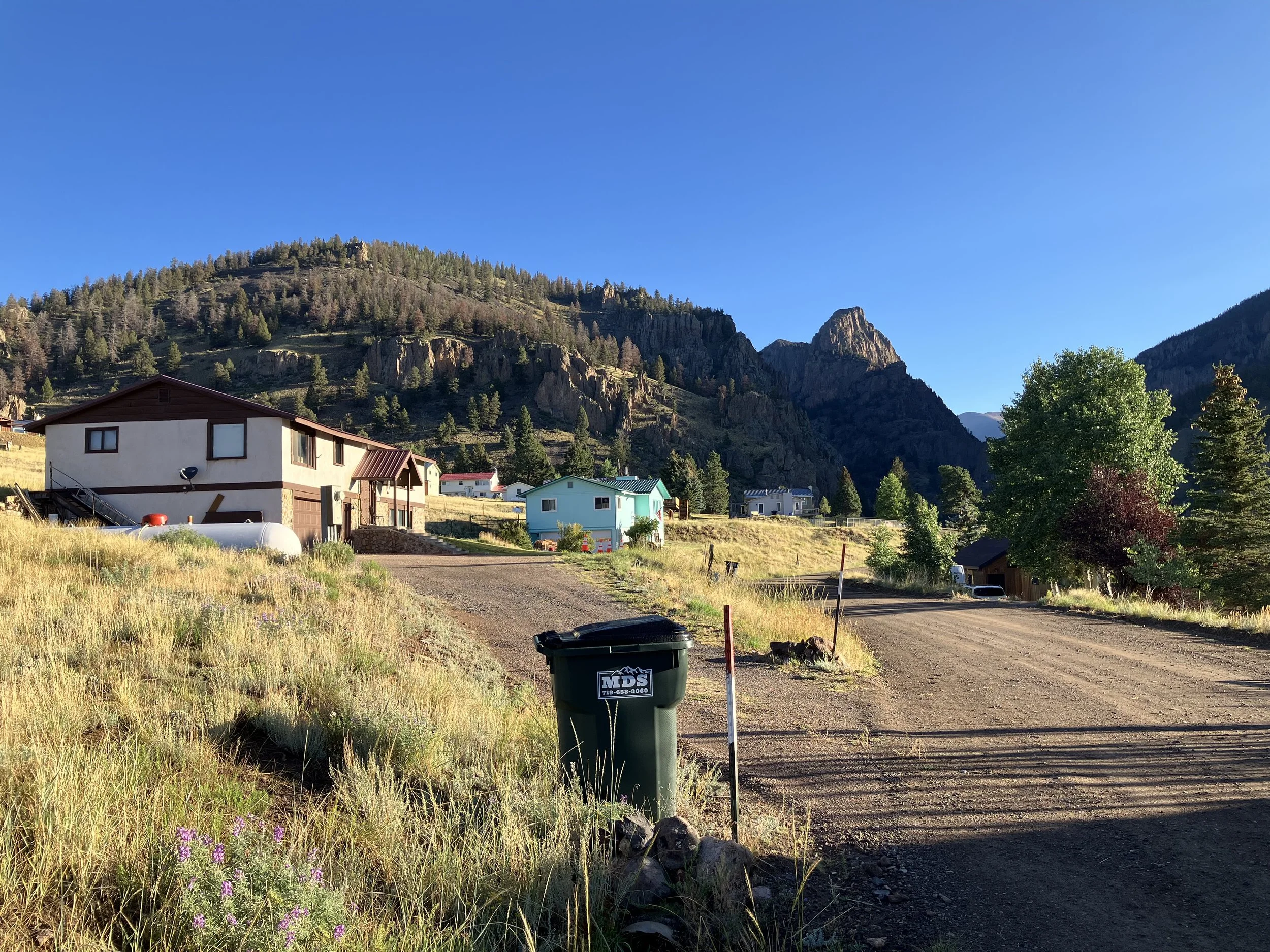 Rural mountain landscape with a dirt road, colorful houses, green trees, and rocky cliffs under a clear blue sky.