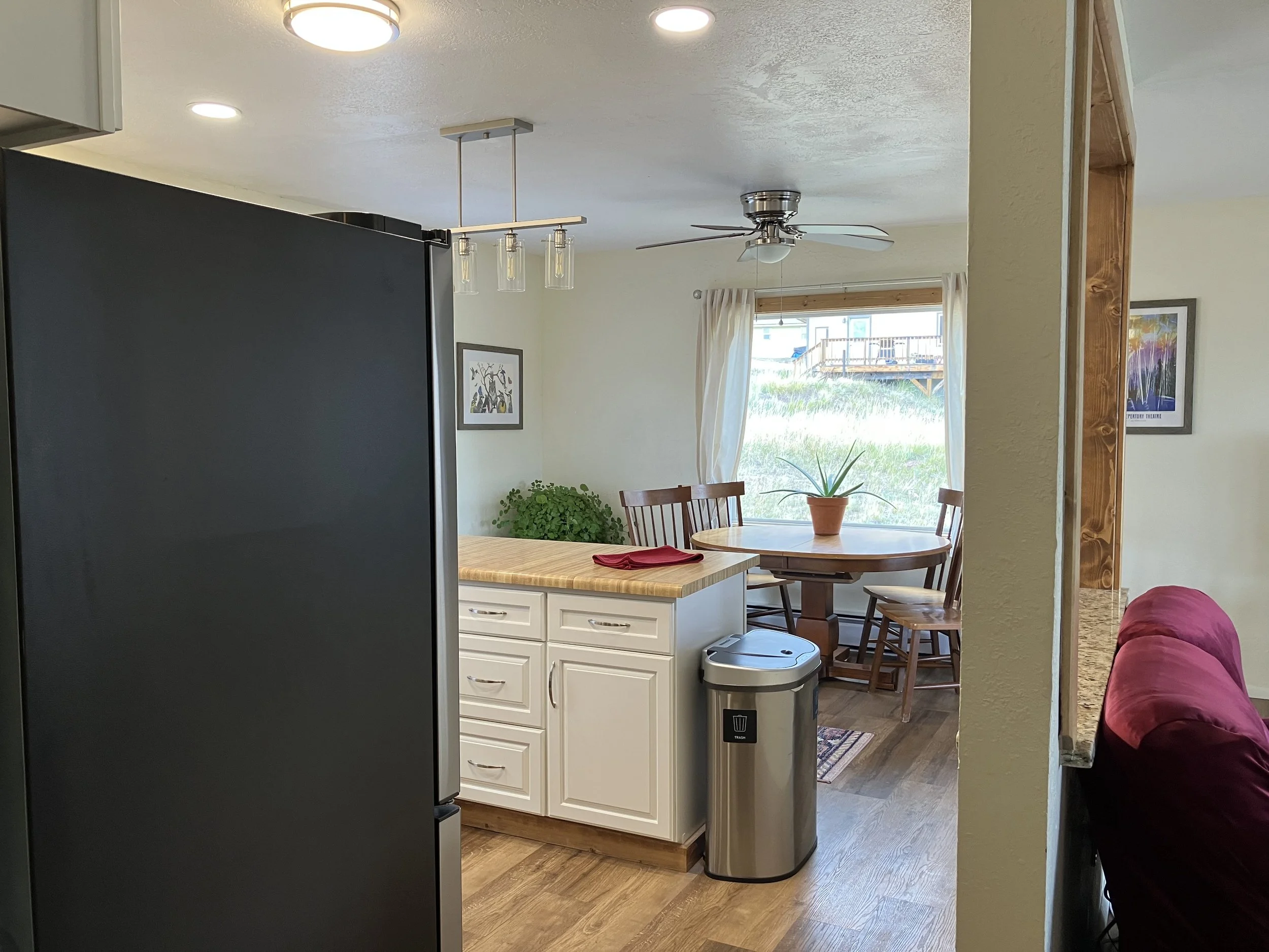 Inside view of a kitchen and dining area with a large window showing an outdoor deck. The kitchen has white cabinets, a wooden countertop, and a trash bin. The dining area has a round wooden table with four chairs and a potted plant. A ceiling fan an