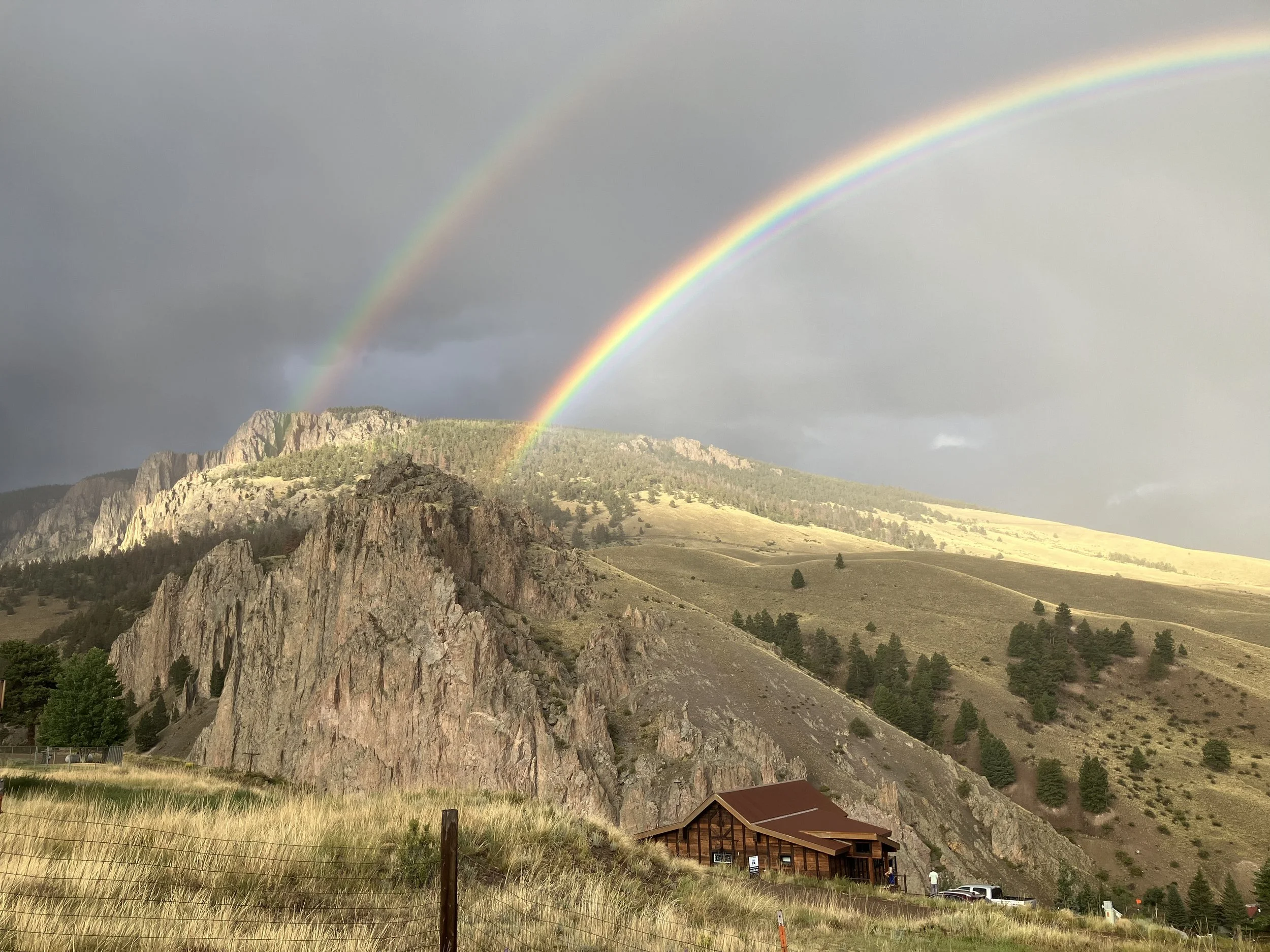 Scenic landscape with double rainbow over rocky mountains, a wooden cabin, and a cloudy sky.