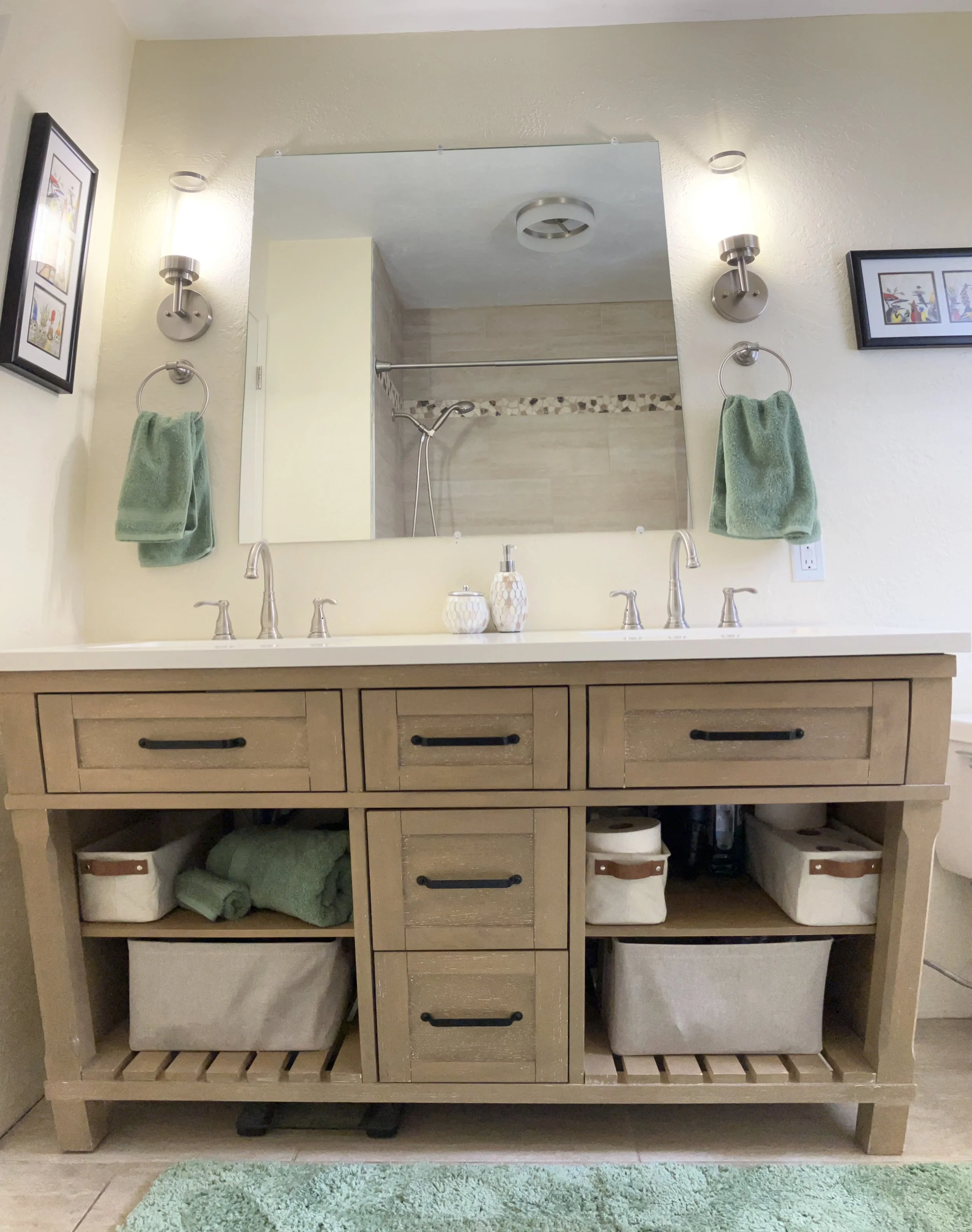 A bathroom vanity with a large mirror, two wall-mounted lights, and two green towels on towel rings. The vanity has six drawers and two open storage compartments with baskets. A turquoise rug is on the floor, and a shower with tile walls and a shower