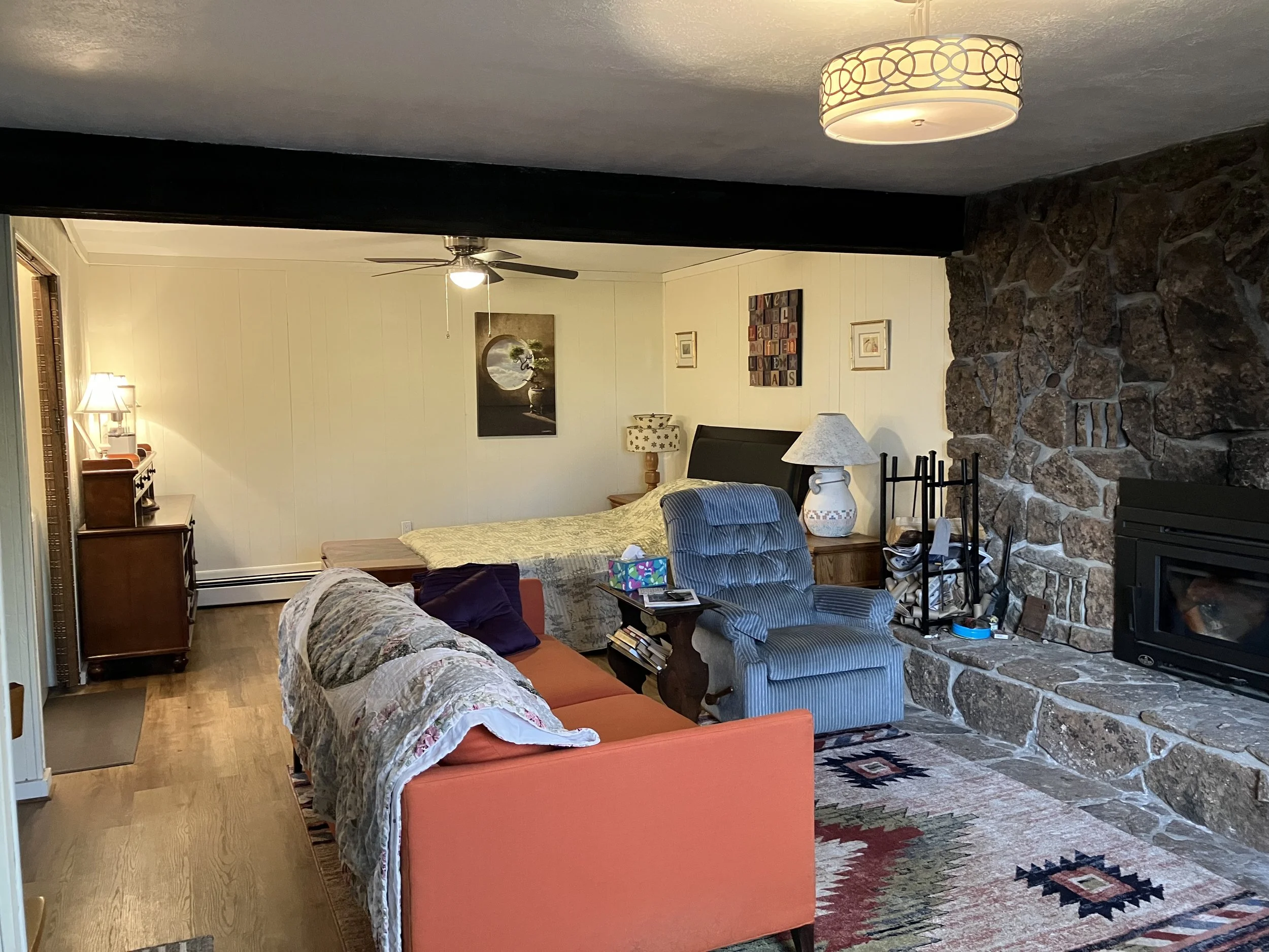 Living room with beige walls and wooden flooring, featuring a stone fireplace on the right, a coral sofa with a quilt on it, a blue striped armchair, a bed in the back, and various side tables and lamps. Decor includes framed artwork and a patterned 
