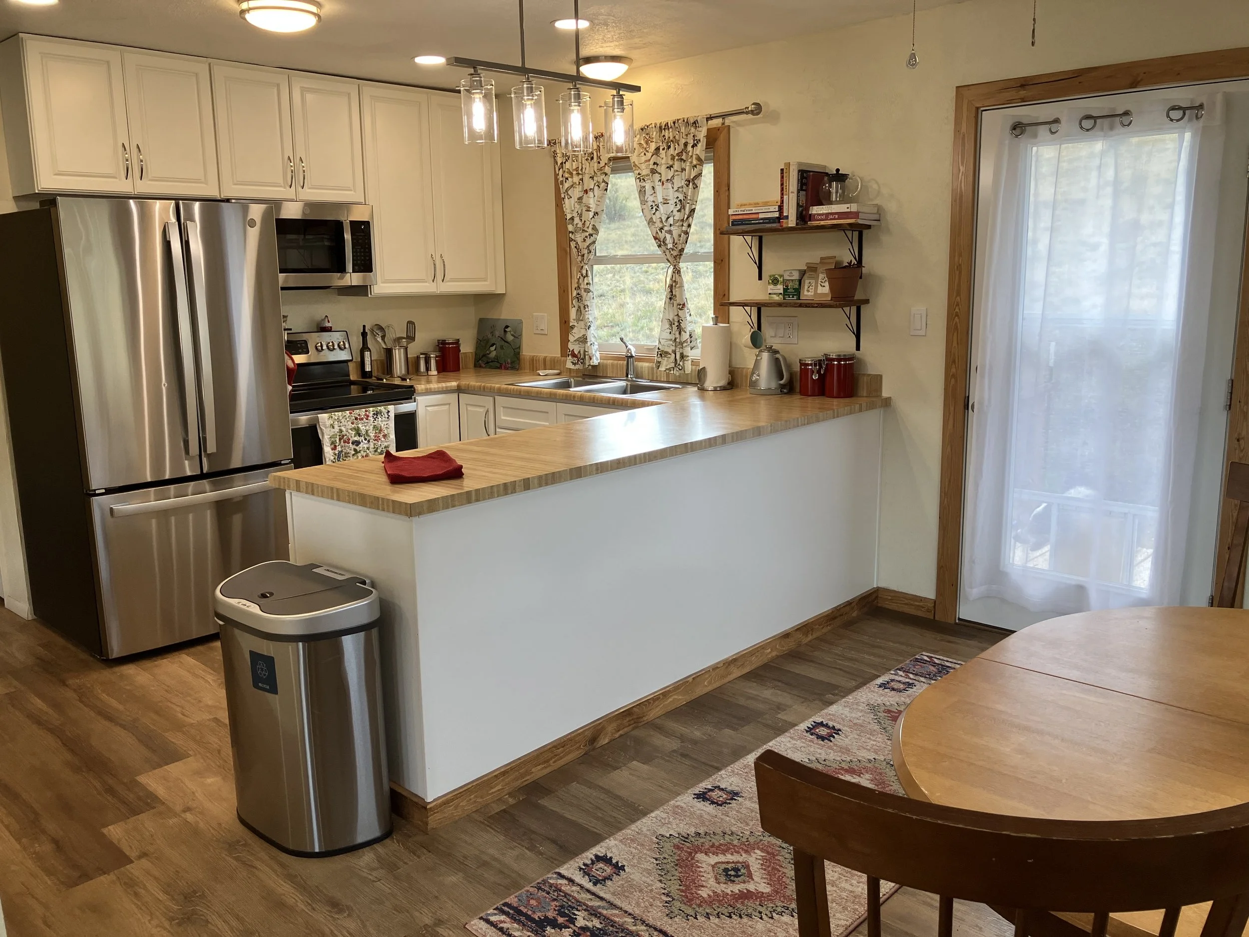 Kitchen with white cabinets, stainless steel refrigerator, oven, microwave, wooden countertop, window with floral curtains, wooden shelves, sliding glass door with curtain, wooden dining table, patterned rug, and trash can.