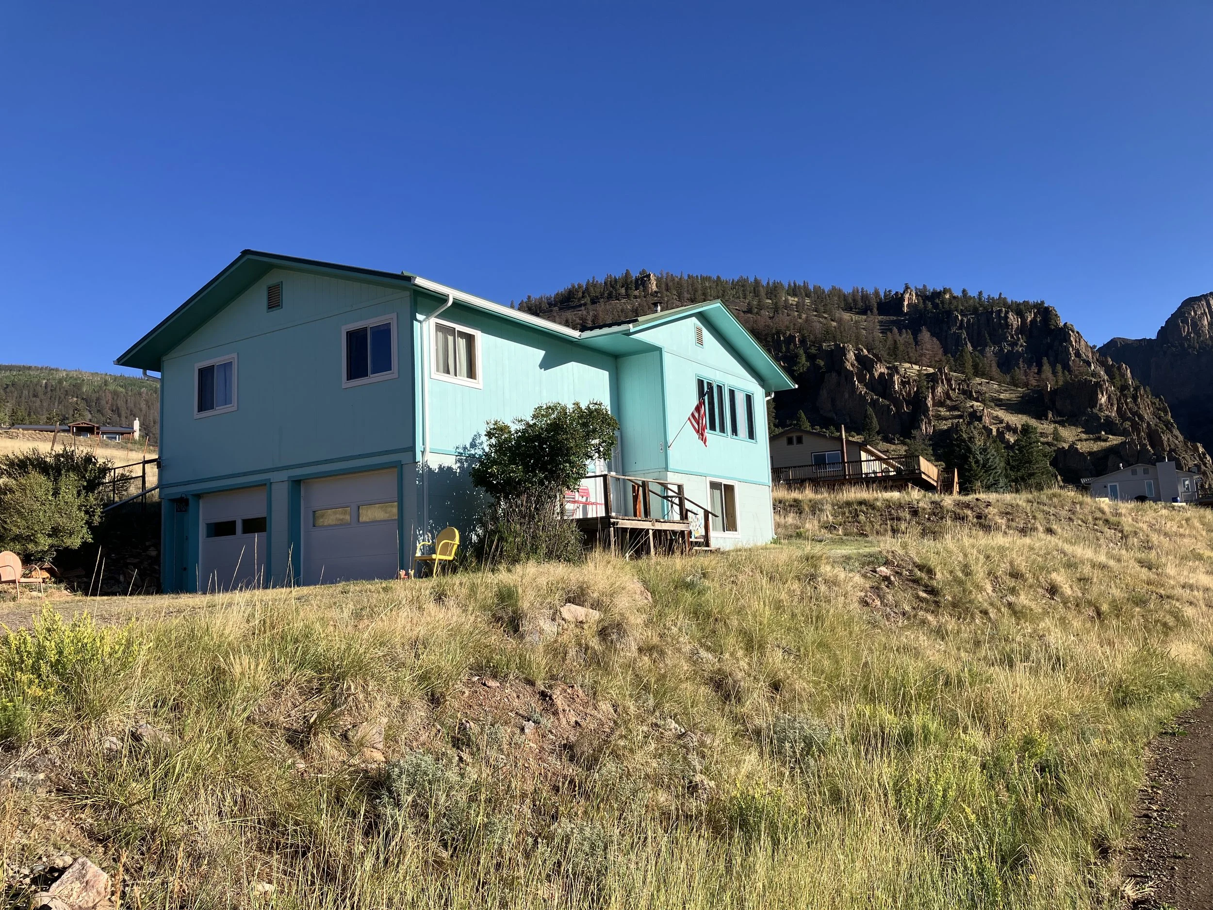 A blue house on a grassy hillside with a mountain and clear blue sky in the background.