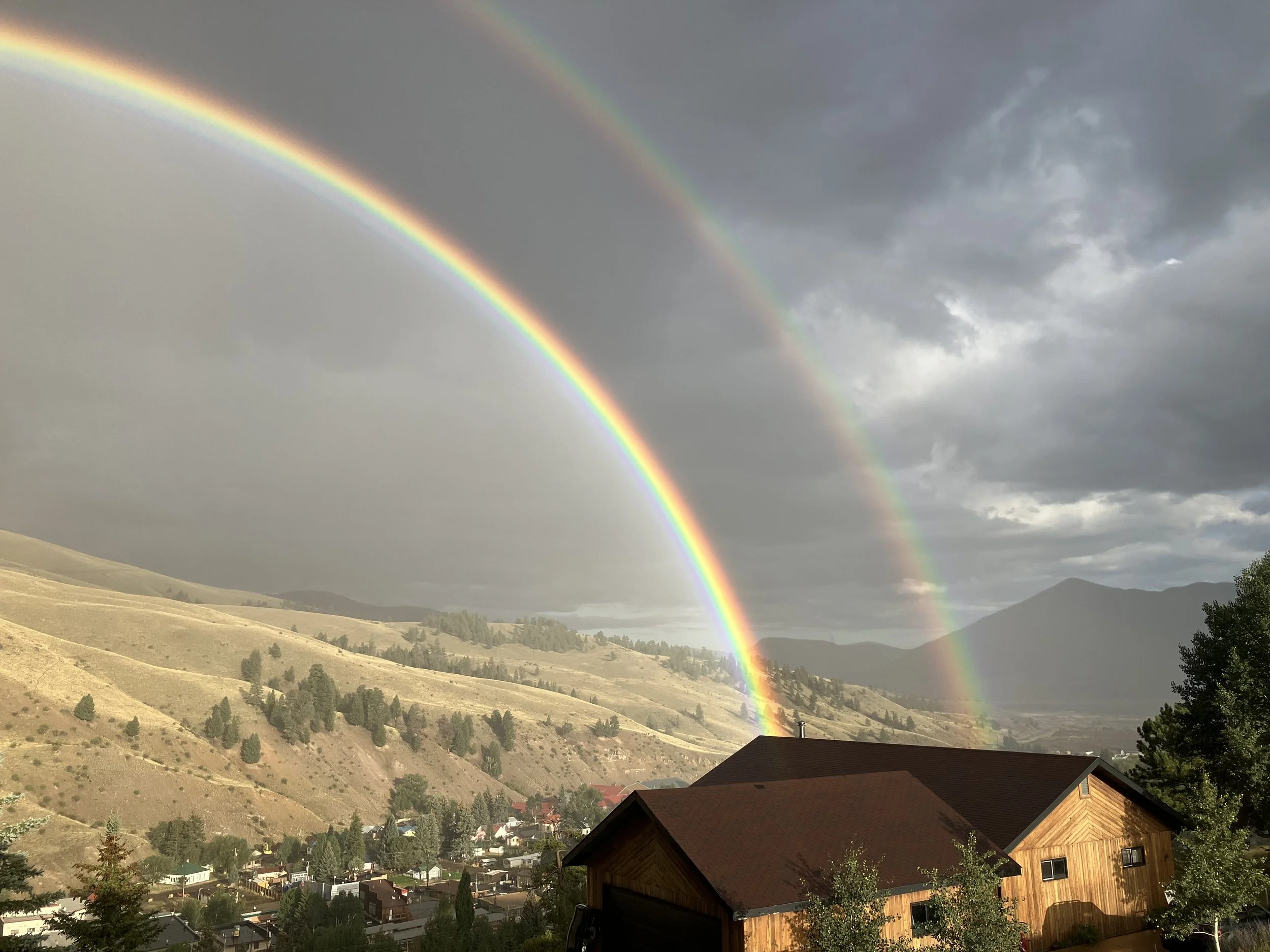 Double rainbow over a hillside town with houses and trees, dark storm clouds in the sky.