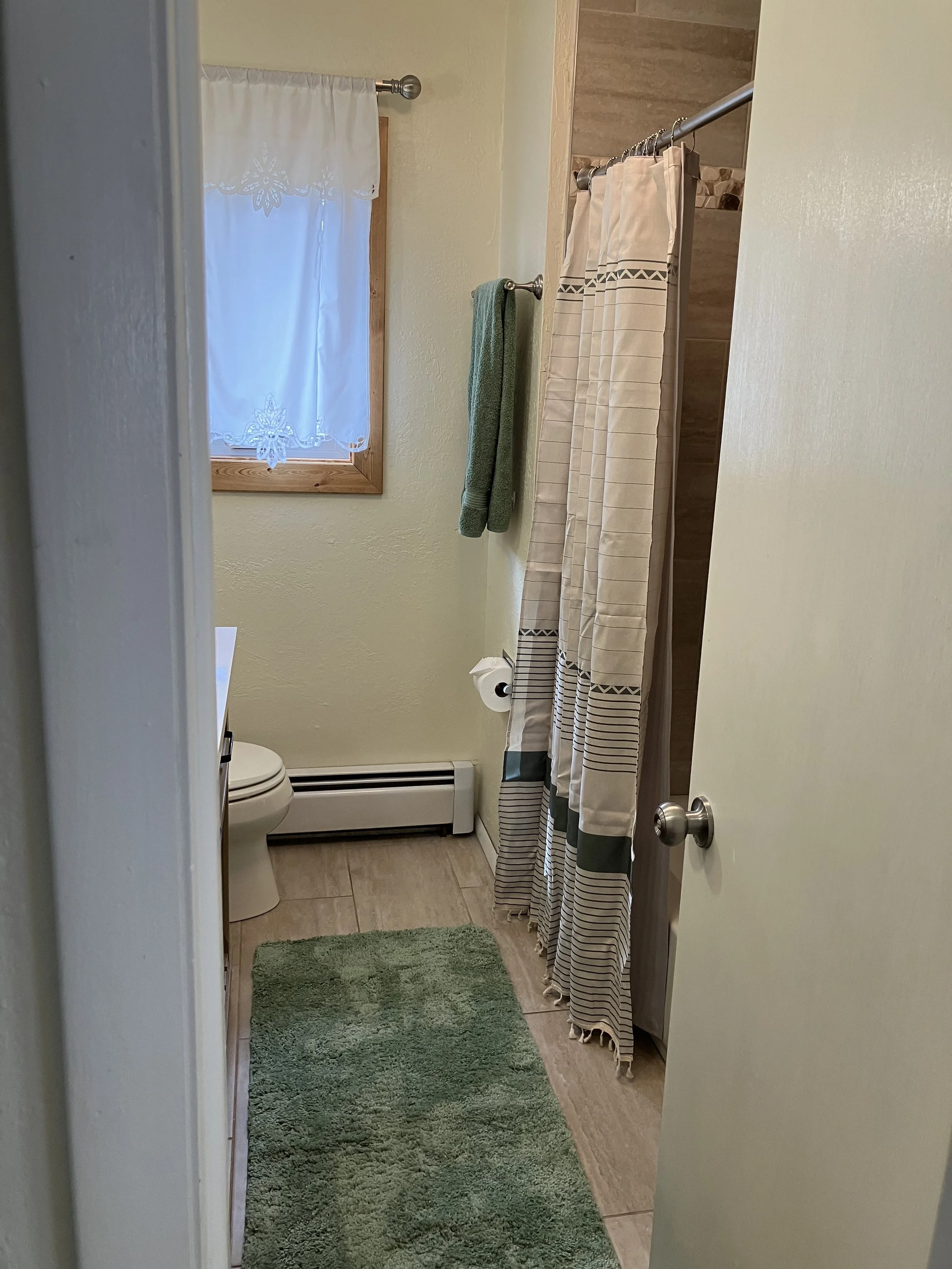 View of a small bathroom with a green rug, a toilet, a window with a white lace curtain, a green towel, and a shower curtain with beige, green, and striped patterns.