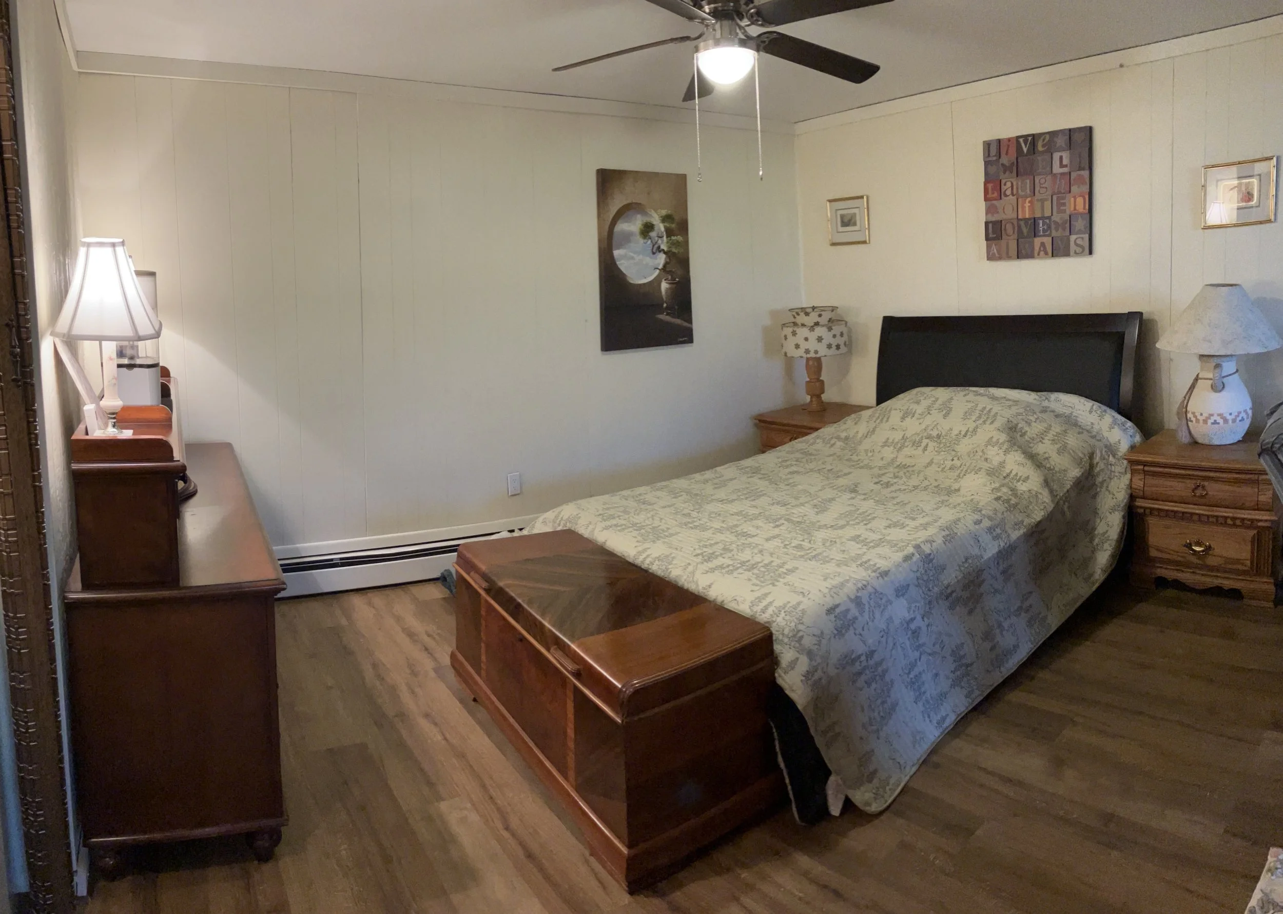 A bedroom with a bed, wooden nightstands, lamps, art on the walls, and a ceiling fan.