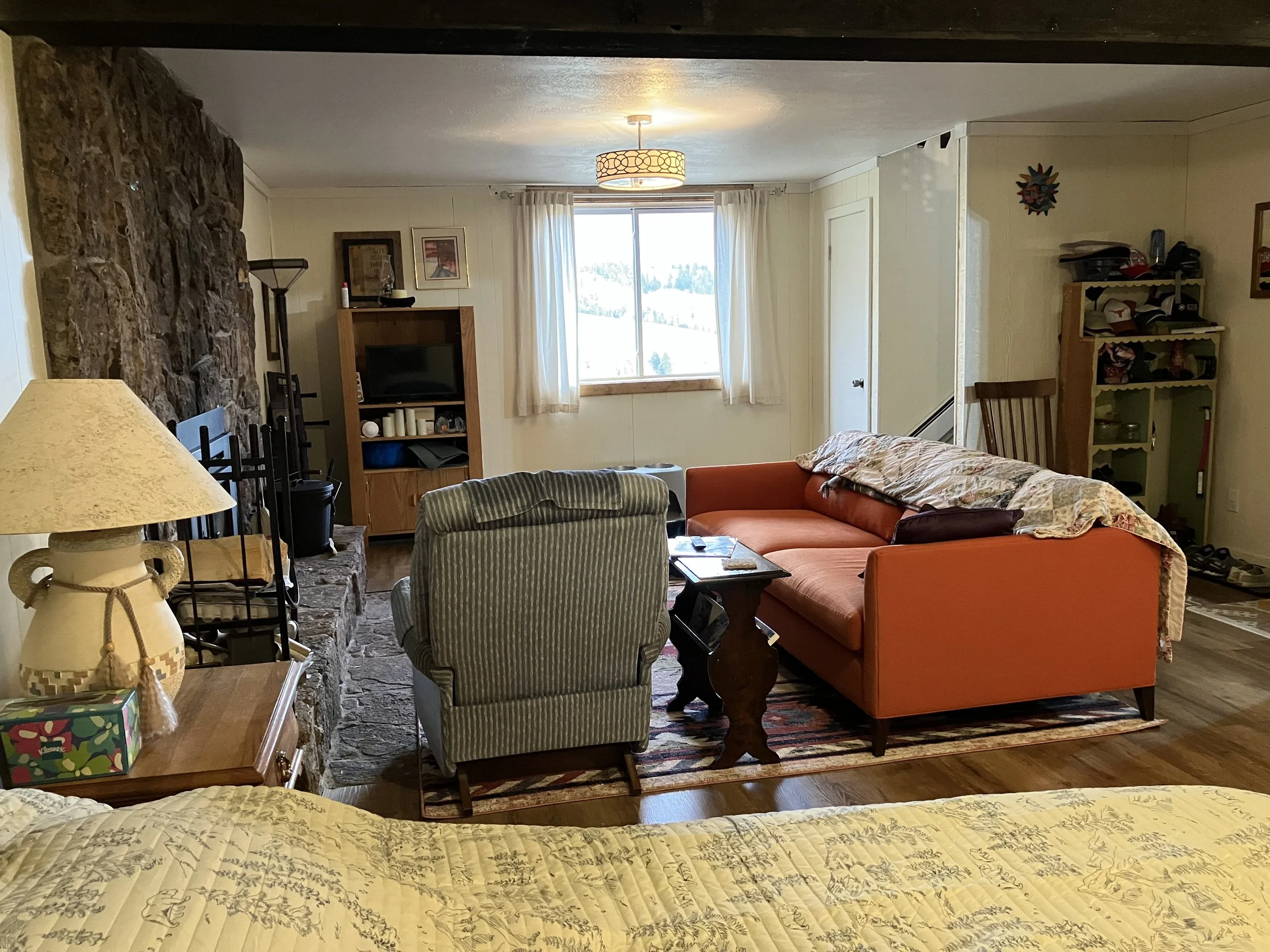 Cozy living room with a stone fireplace, an orange sofa covered with a quilt, a green striped armchair, a small wooden coffee table, a TV on a wooden stand, and a window with white curtains allowing natural light.