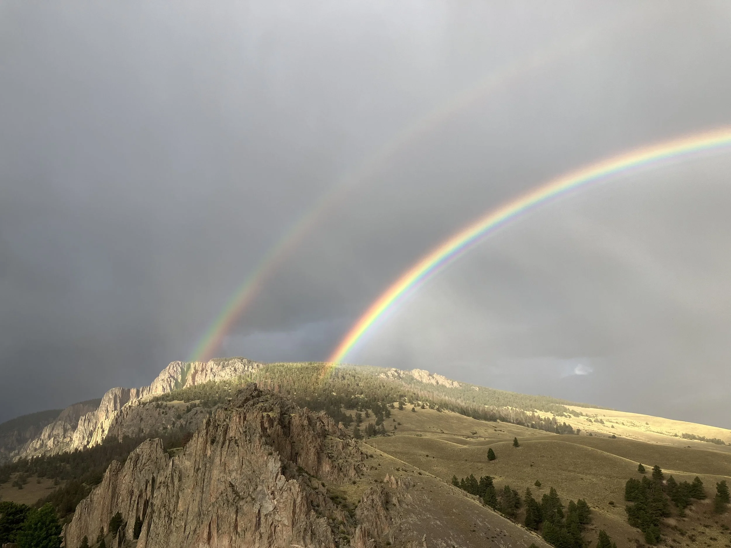 Two rainbows appear in a cloudy sky over a rugged mountain landscape with a grassy hillside and sparse trees.