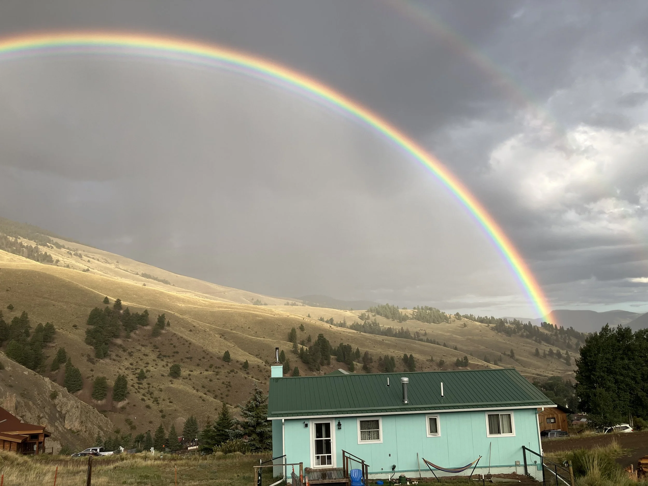 A landscape with a rainbow arching over rolling hills and a turquoise house in the foreground.