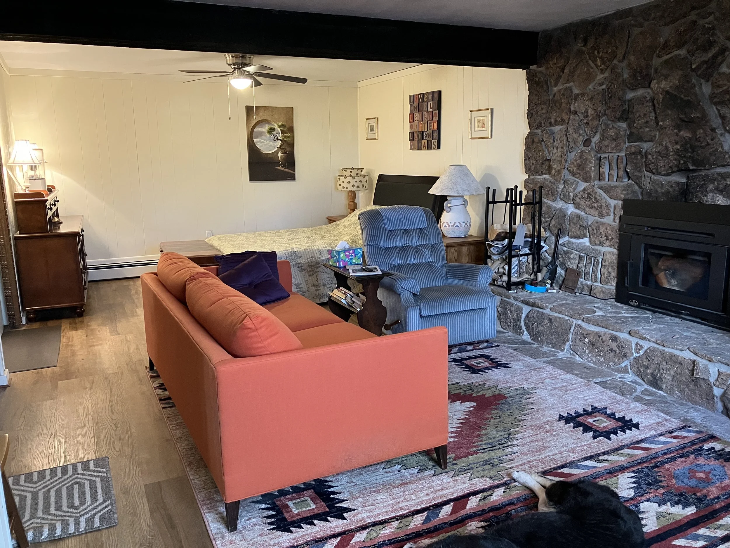 Living room with a burnt orange sofa, a blue-striped armchair, a bed in the background, a stone fireplace, area rugs, and various decorative items and furniture.