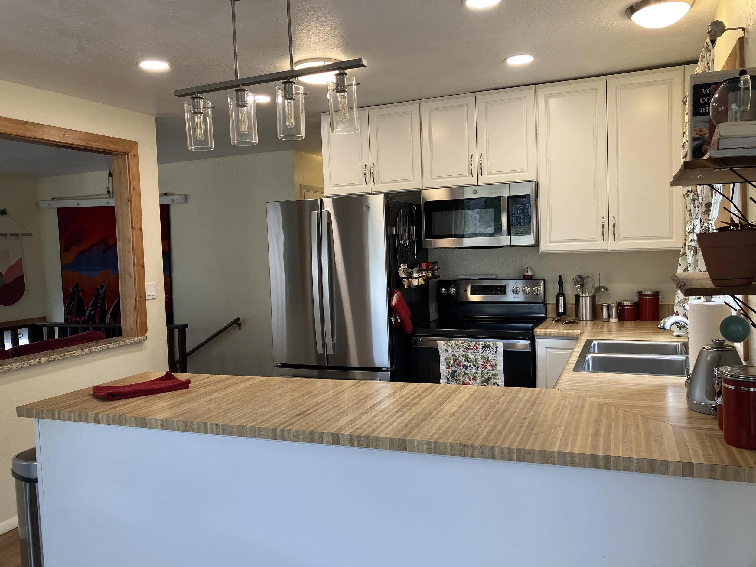 Kitchen with white cabinets, stainless steel appliances including refrigerator and microwave, wooden countertops, and a window with floral curtains.