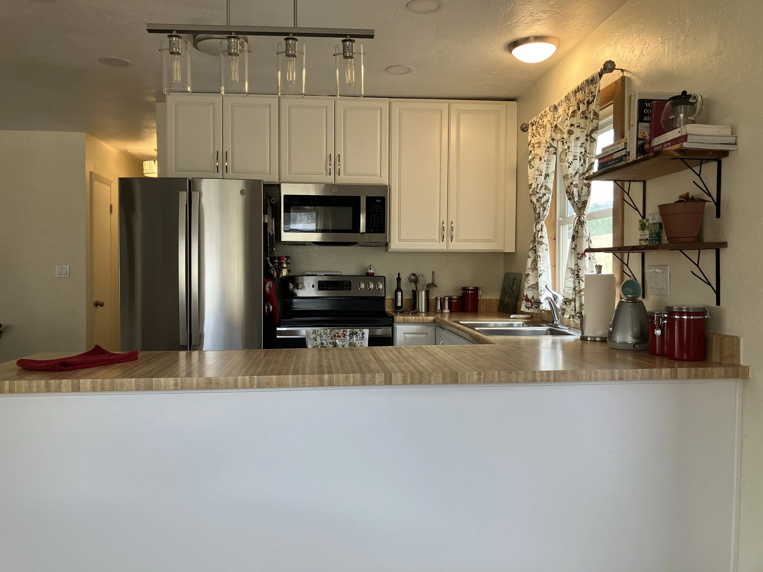 Kitchen with white cabinets, stainless steel refrigerator, microwave, and oven; wooden countertop with red canisters and kitchen appliances; window with patterned curtains; wood shelves with books, plants, and kitchen items.