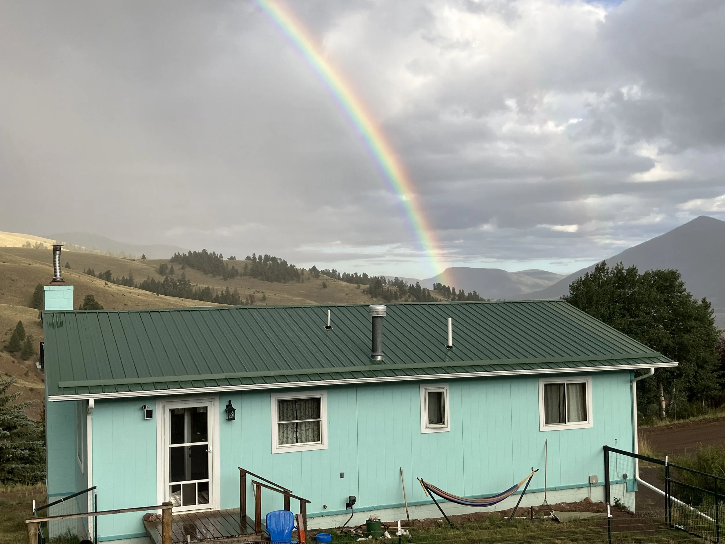 A turquoise house with a green metal roof in a mountainous area. A rainbow arches across a cloudy sky in the background.