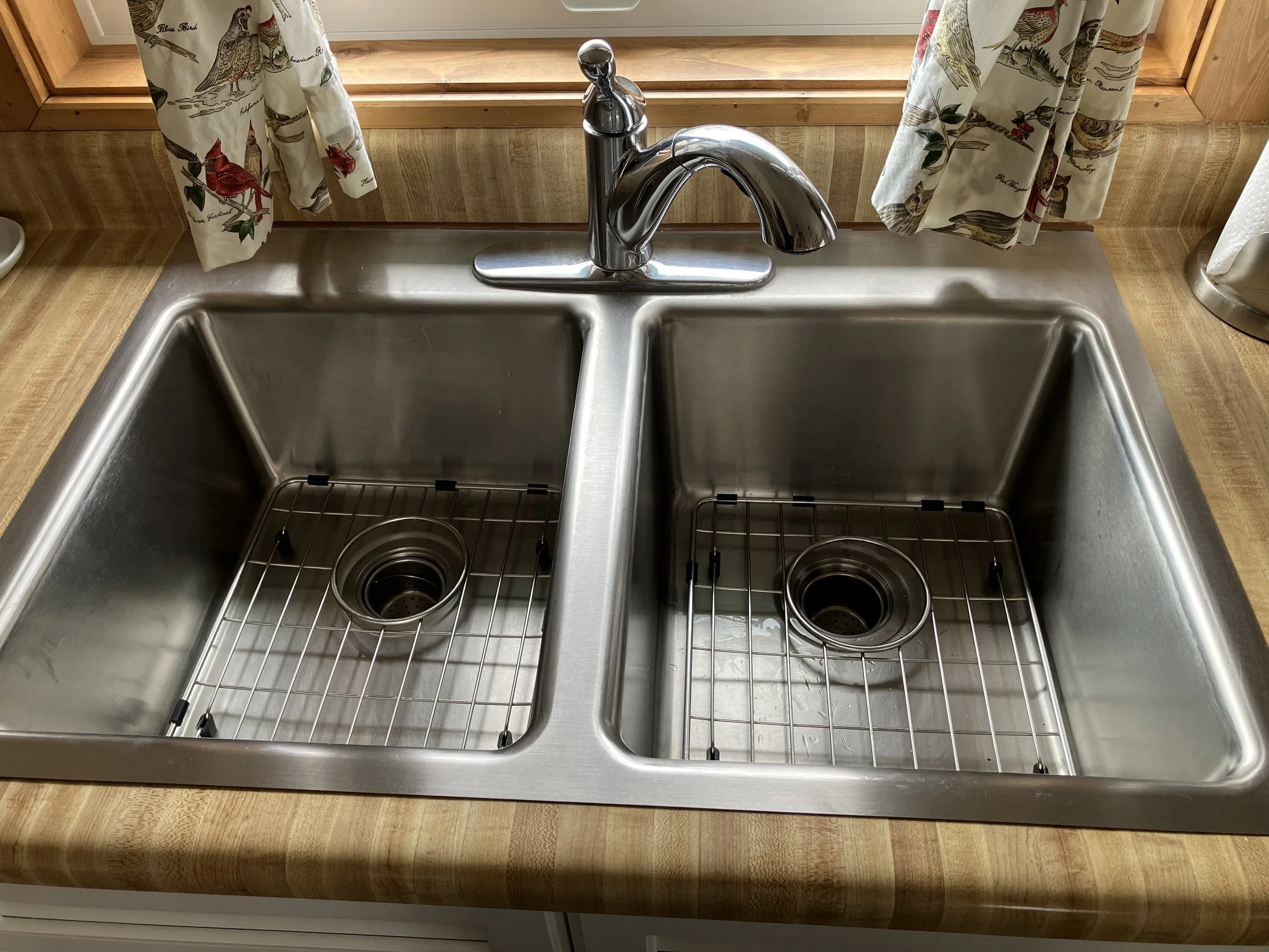 Double stainless steel kitchen sink with two basins, each with a removable wire rack at the bottom, and a chrome faucet centered above the divider. The sink is installed in a wooden countertop with a window and patterned curtains in the background.