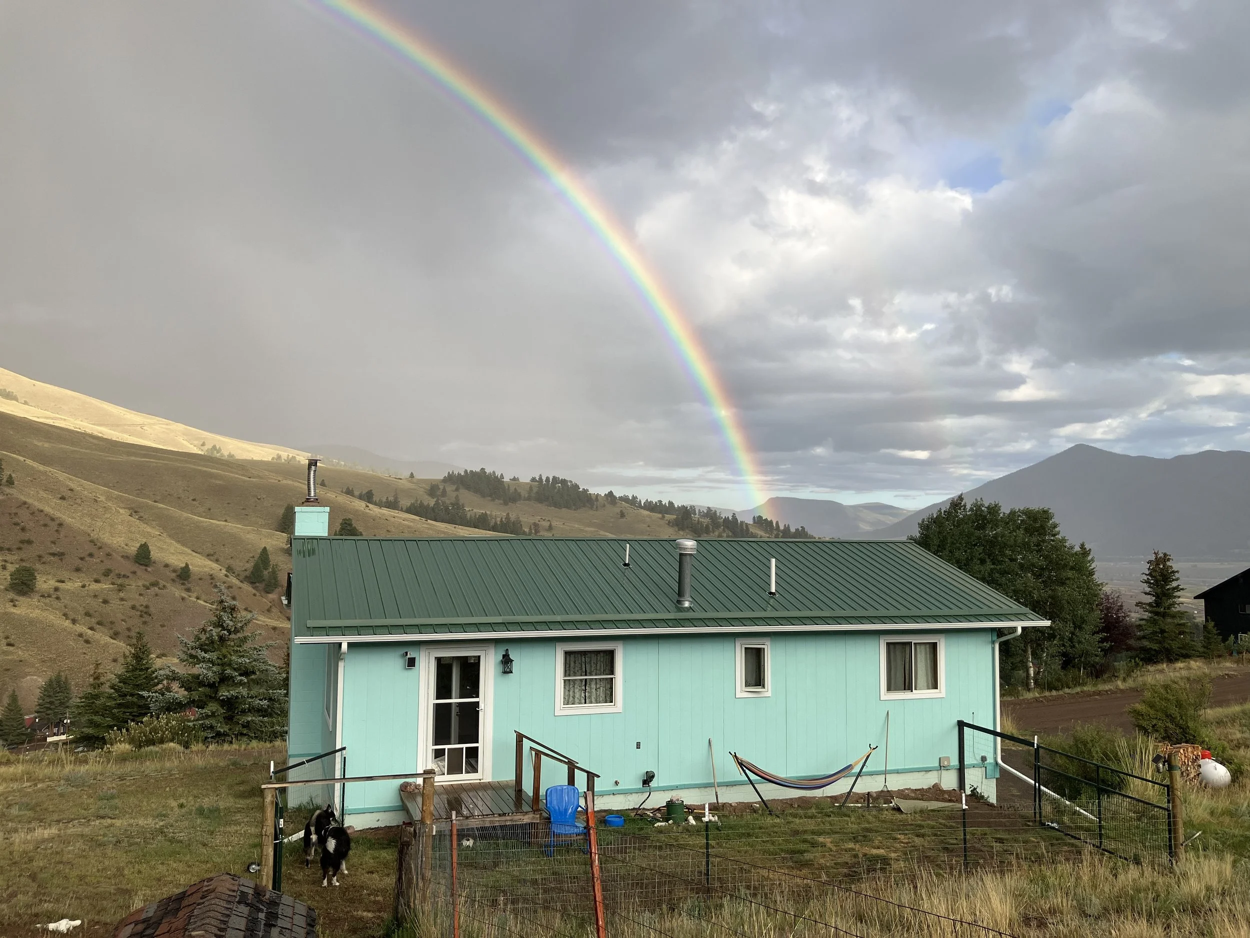 A turquoise house with a green metal roof in a rural landscape under a cloudy sky with a rainbow.