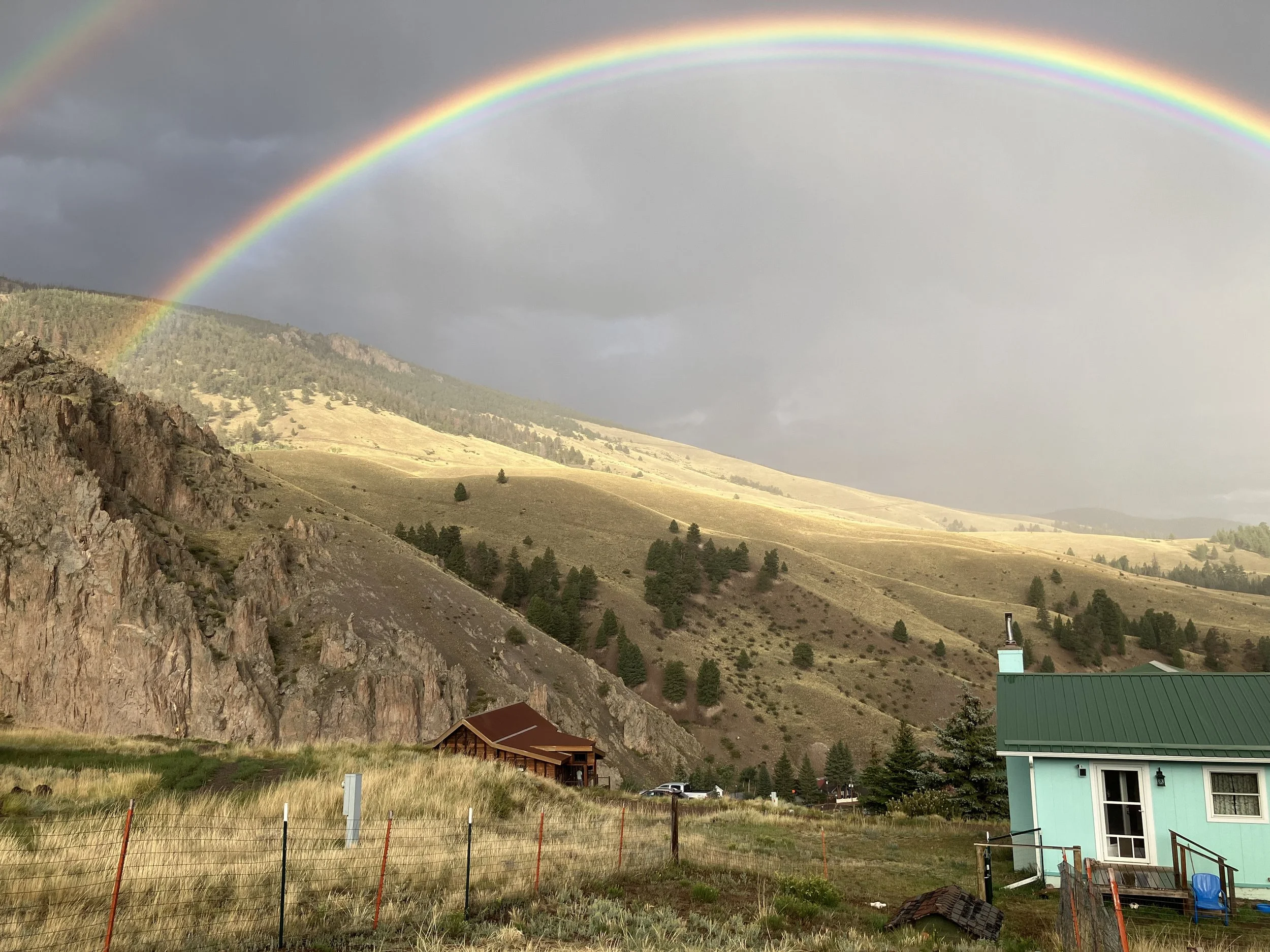 Rainbow over a hilly landscape with a turquoise house in the foreground, a mountain with rocky outcrops on the left, and a partly cloudy sky.