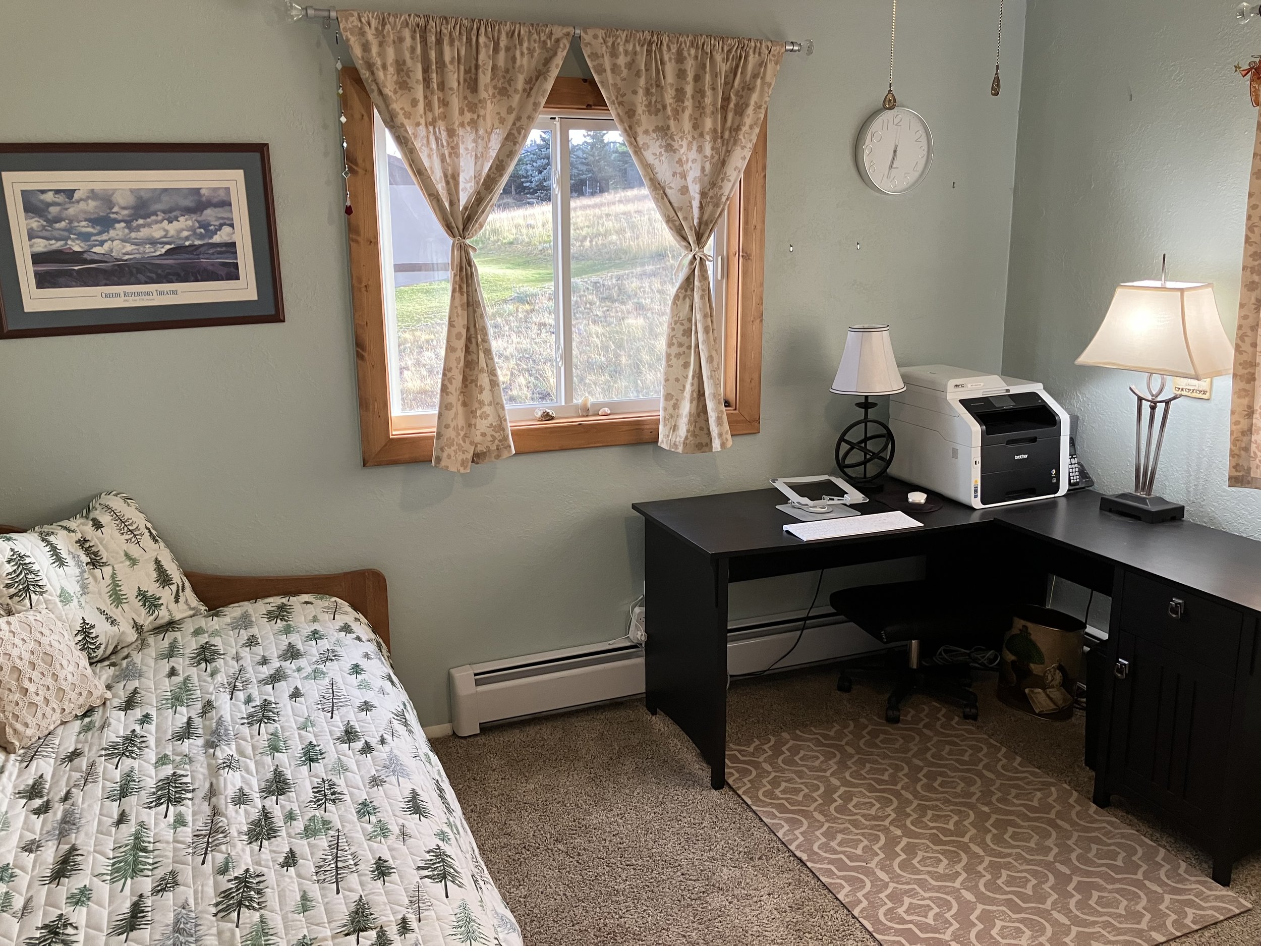 A cozy bedroom corner with a bed covered in a pine tree patterned quilt and a decorative pillow, a window with beige floral curtains letting in natural light, a black desk with a white printer, a lamp, papers, and a phone, a black chair, a beige patt