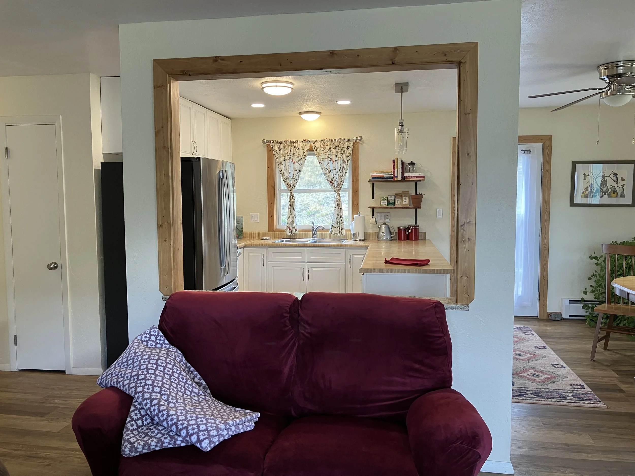 Living room with a view into the kitchen, visible are a burgundy couch with a patterned pillow, hardwood floors, and a kitchen window with floral curtains, light-colored cabinets, and a wooden framed opening between the two rooms.