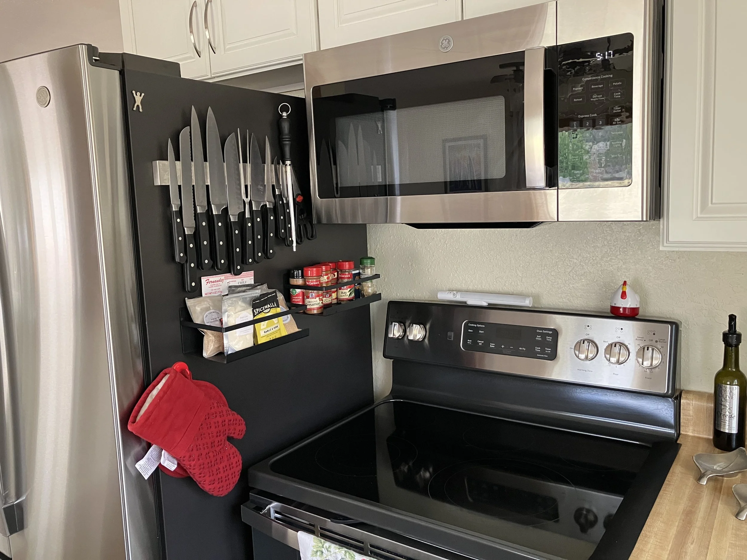 Kitchen with a stainless steel microwave above a black electric stove, a magnetic knife holder with knives, scissors, and food spices, a red oven mitt hanging on the side of the black panel, a bottle of olive oil on the countertop, and a small red an