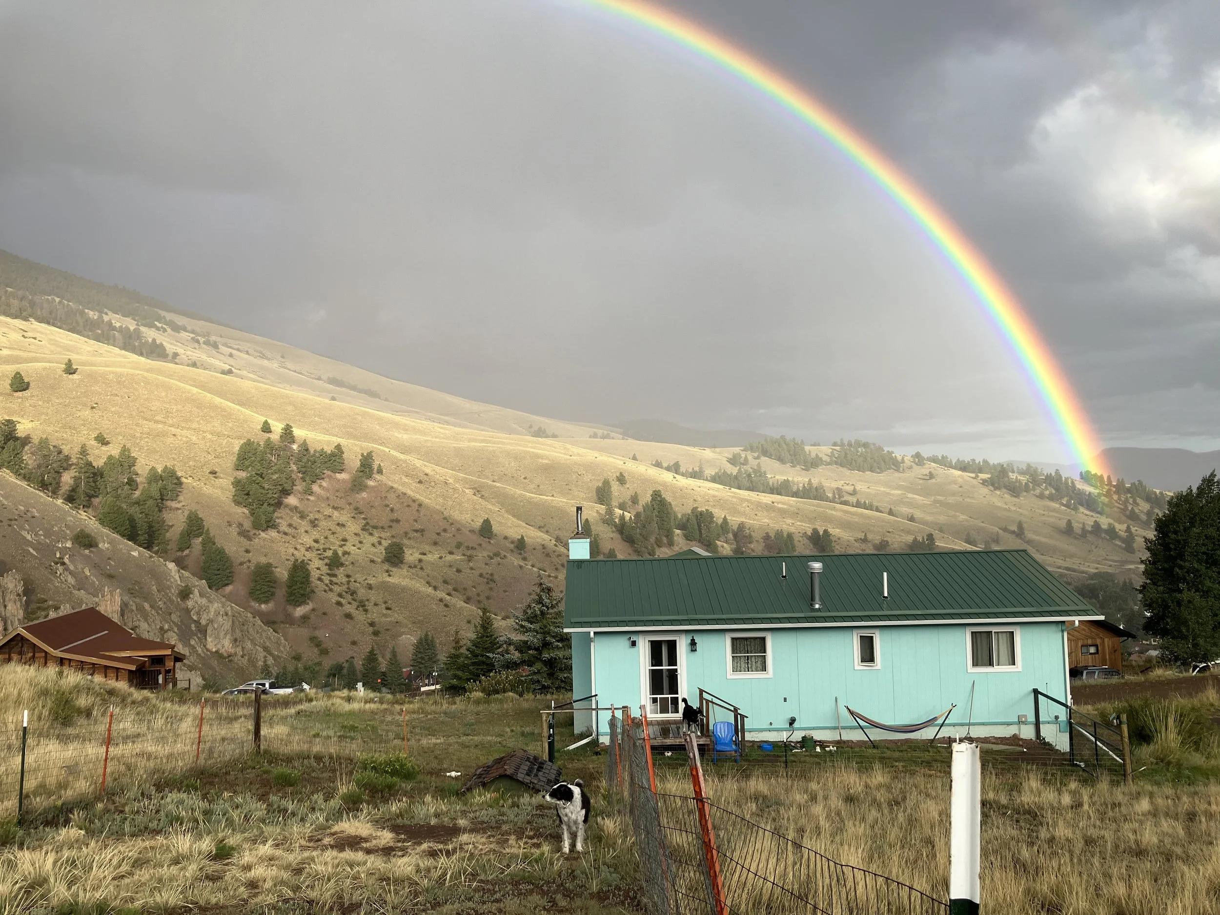 A light blue house with a green roof under a rainbow in a rural landscape with hills and trees.