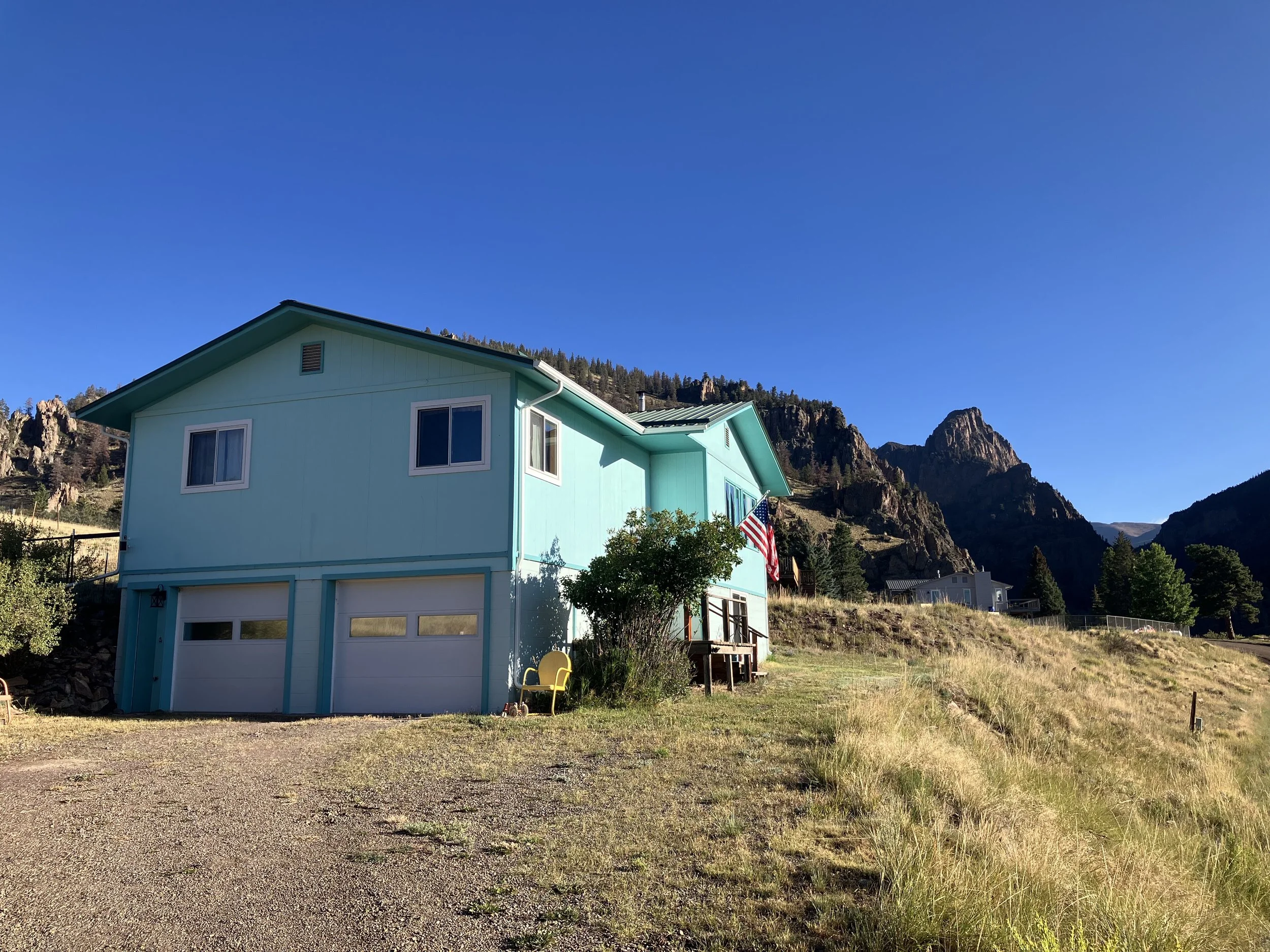 A turquoise two-story house with a garage, set in a hilly landscape with rocky mountains and a clear blue sky in the background.