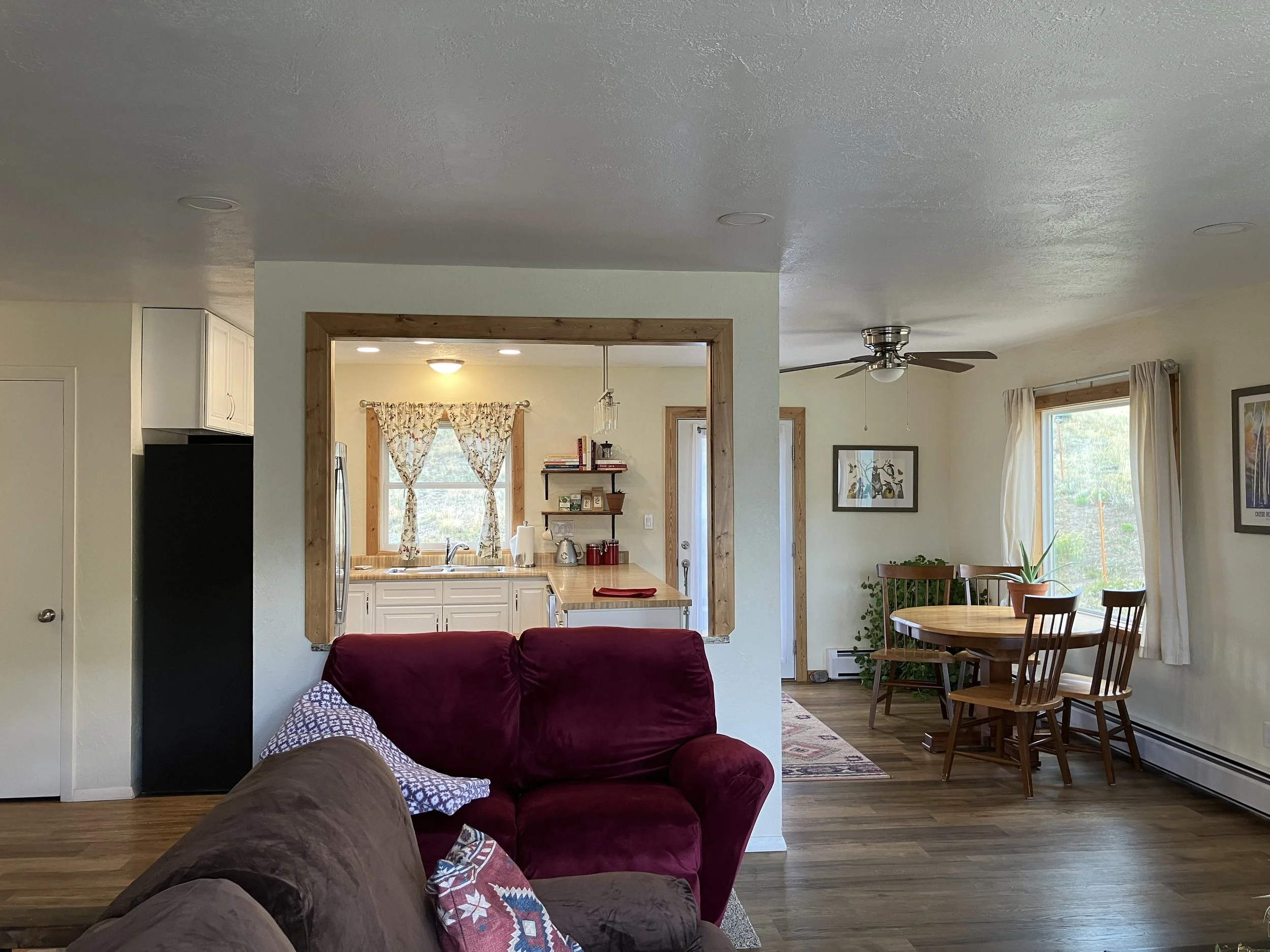 Living room opening into a kitchen and dining area with a red armchair, a brown sofa, a round wooden dining table with four chairs, large windows with white curtains, hardwood floors, a ceiling fan, and decor on the walls and shelves.