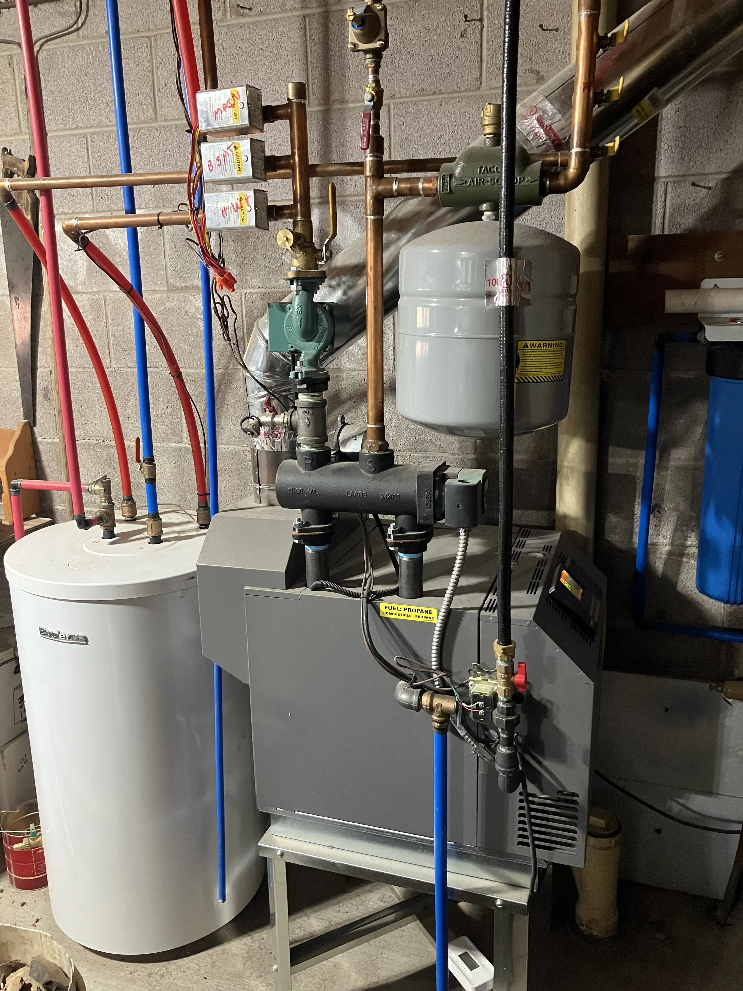 This image shows a boiler system with various pipes, valves, and gauges installed on a concrete wall in a utility room.