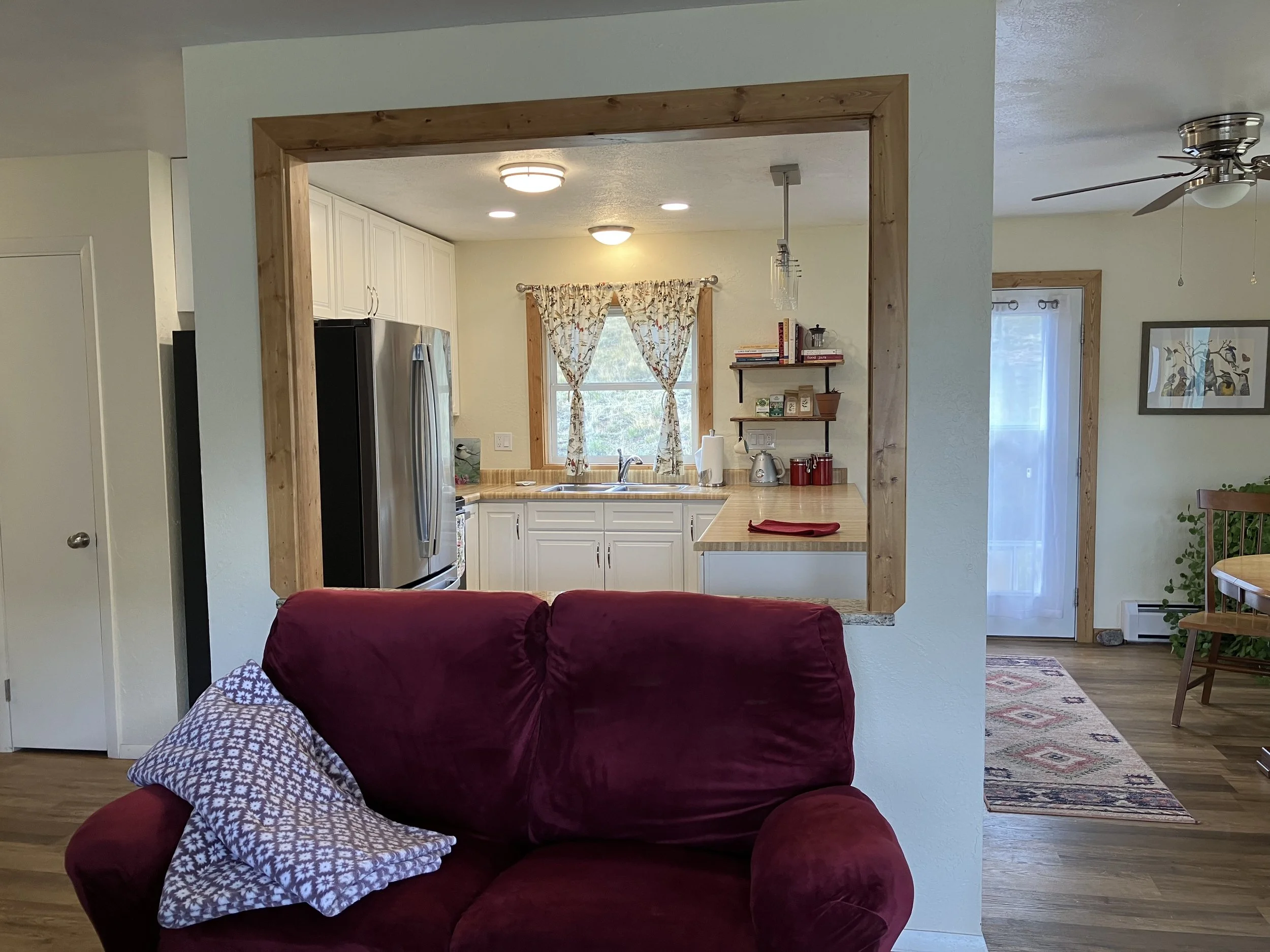 View of a kitchen seen through a wall opening from a living room with a red upholstered couch. The kitchen has white cabinets, a stainless steel refrigerator, a window with patterned curtains, wooden shelving with books and appliances, and ceiling li