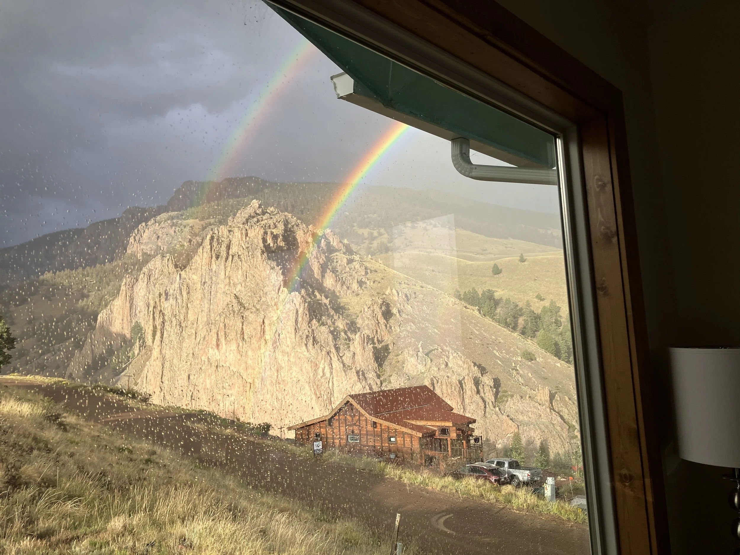 View of a mountain with a double rainbow outside a window on a rainy day.