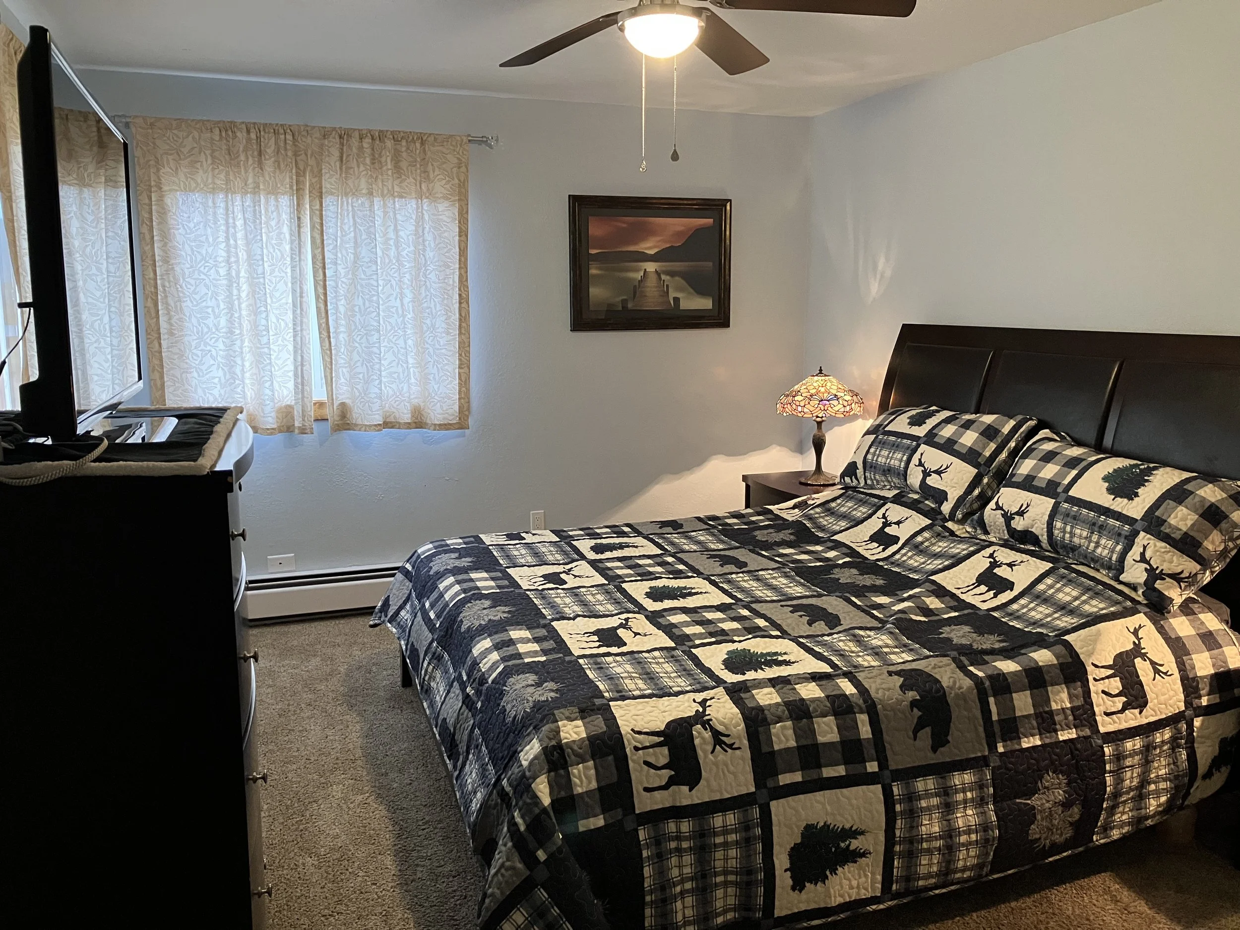 A bedroom with a bed covered in a plaid quilt featuring moose, trees, and bears, a wooden nightstand with a stained glass lamp, a dresser with a television, and a window with beige curtains.