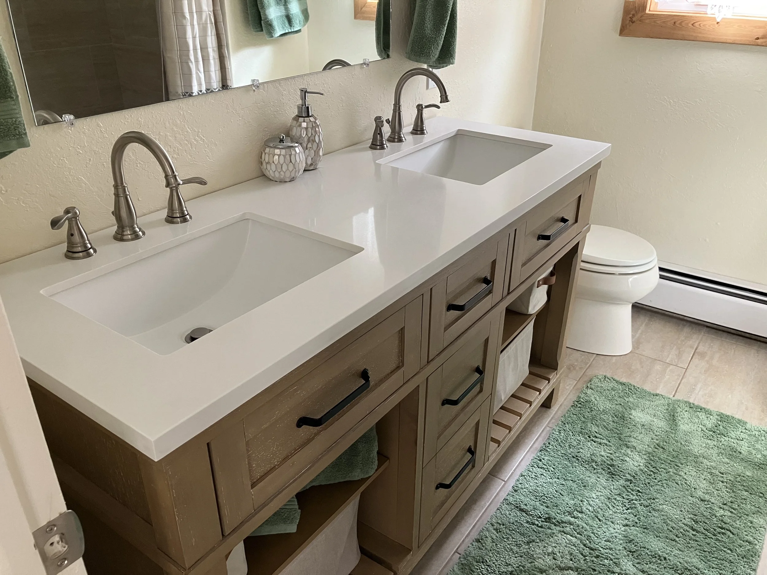 A bathroom with a double vanity featuring a white countertop, two sinks, and modern brushed nickel faucets. There are matching soap dispensers and a mirror above the vanity. To the right, a toilet is visible, and a green towel hangs on a towel rack a