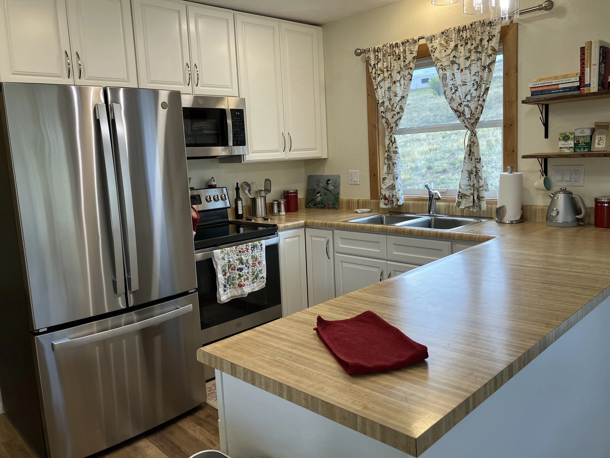 Kitchen with white cabinets, stainless steel refrigerator, microwave, and stove. Wooden countertops, a window with floral curtains, and various small appliances on the counter.