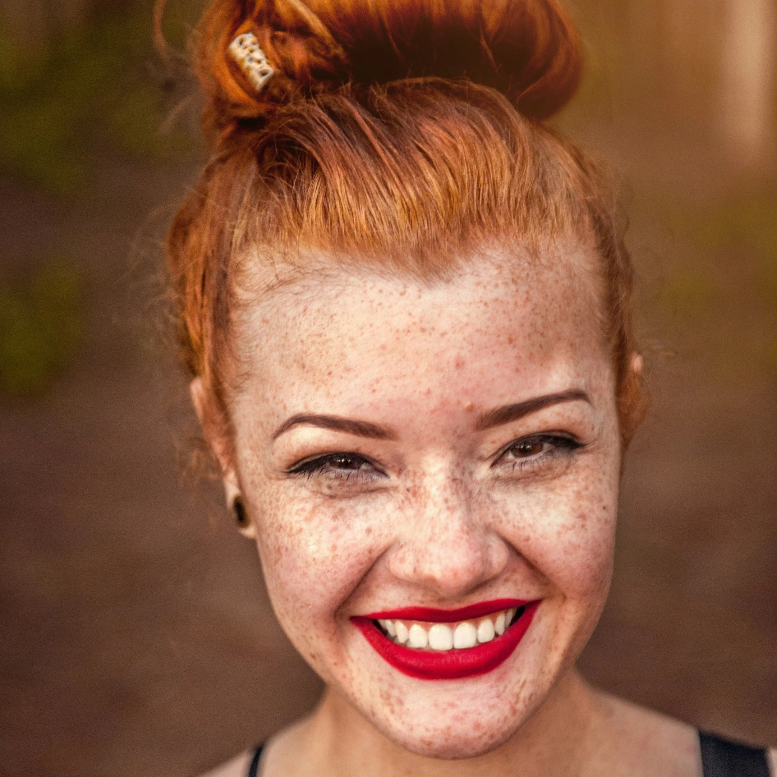 Close-up of a smiling young woman with red hair, freckles, and bright red lipstick, outdoors with a blurred background.