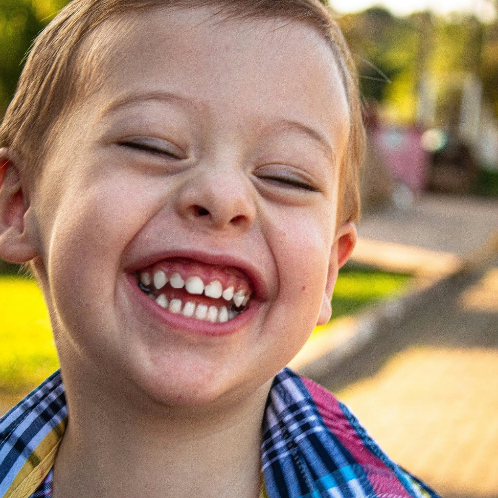 Close-up of smiling young boy with missing teeth, wearing a checkered shirt, outdoors in a park or street.