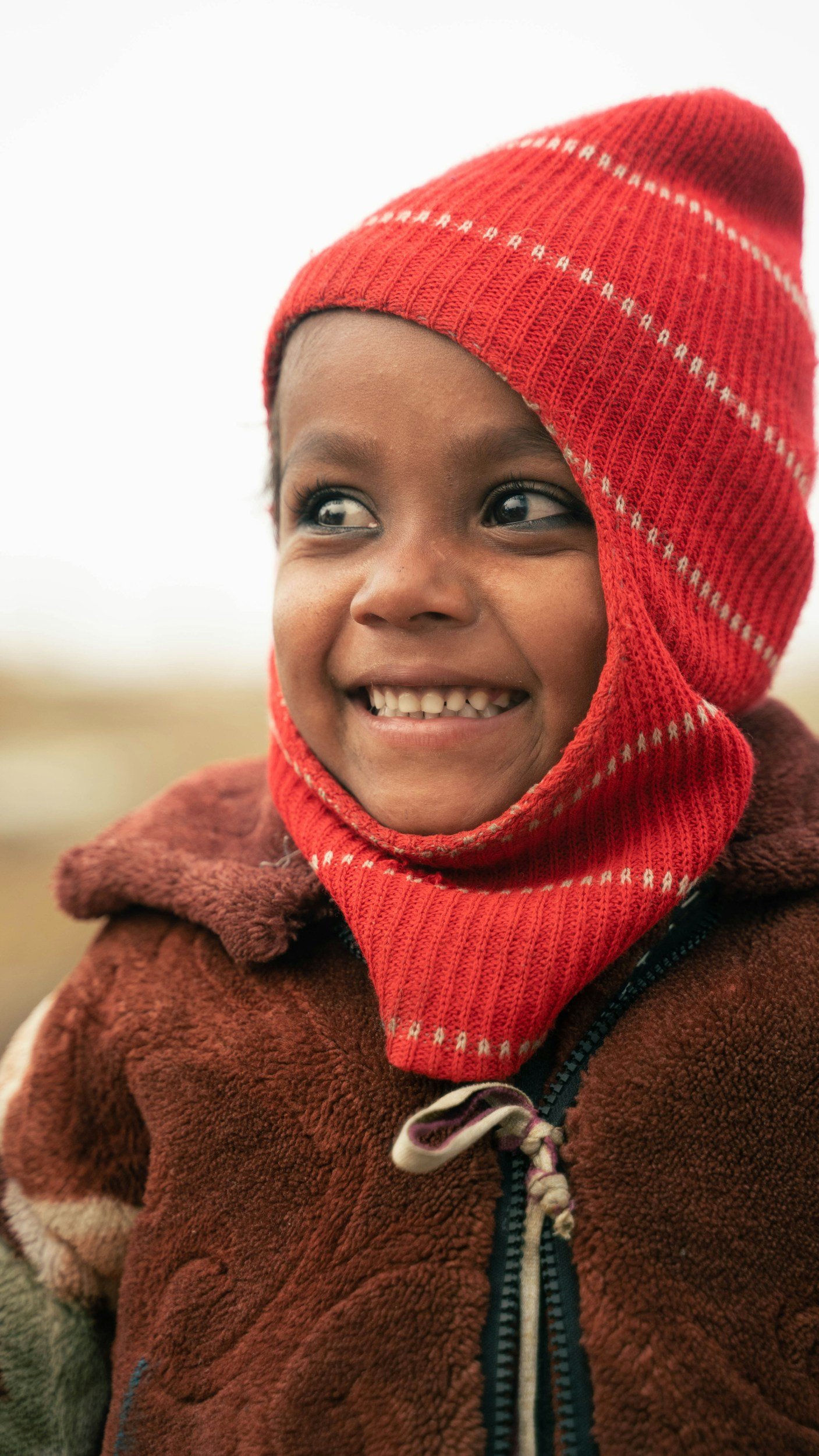 A young child smiling outdoors, wearing a red knit hat and brown fleece jacket.