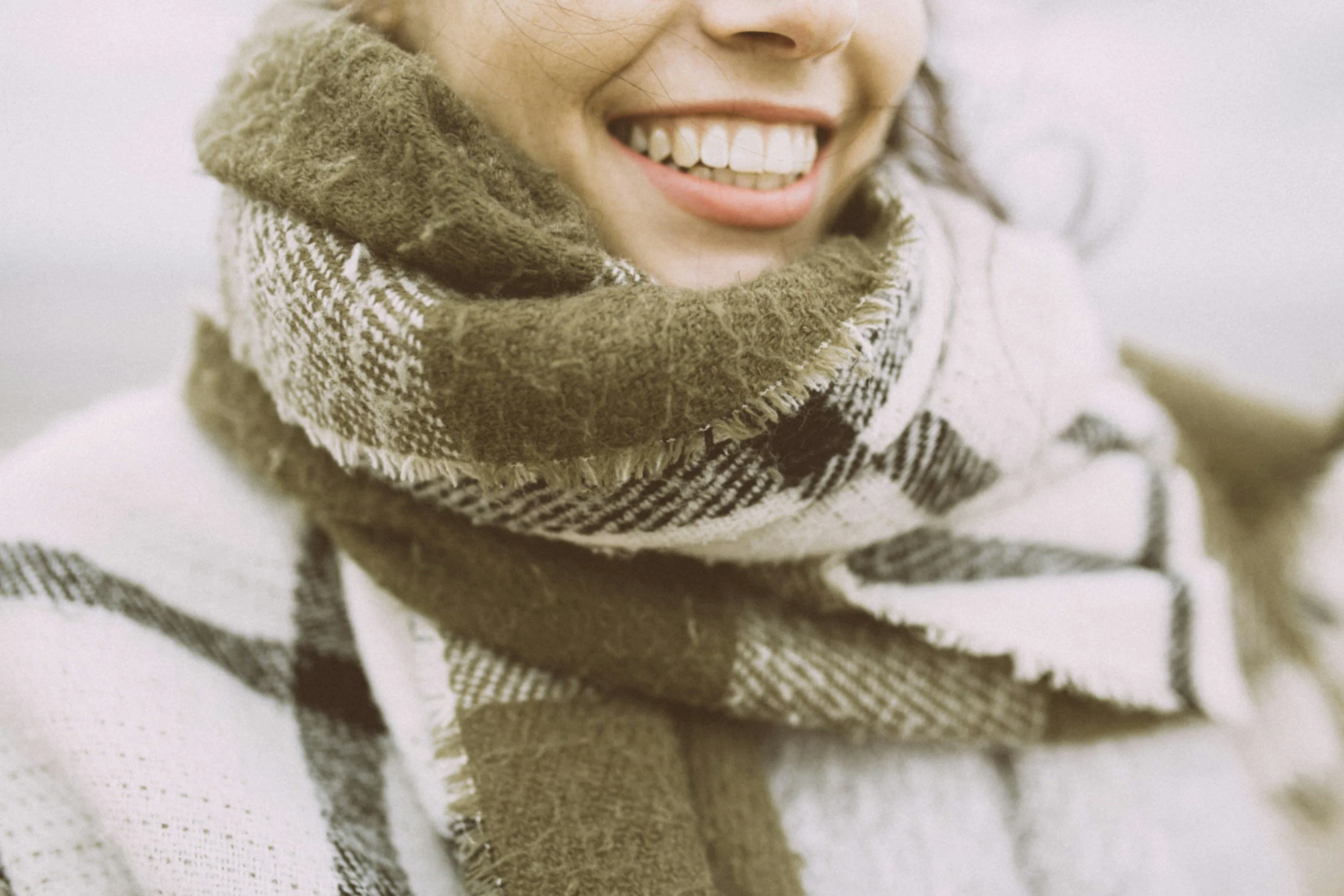 Close-up of a woman smiling, wearing a brown and white checkered scarf with fringed edges, and a white coat with black plaid patterns.