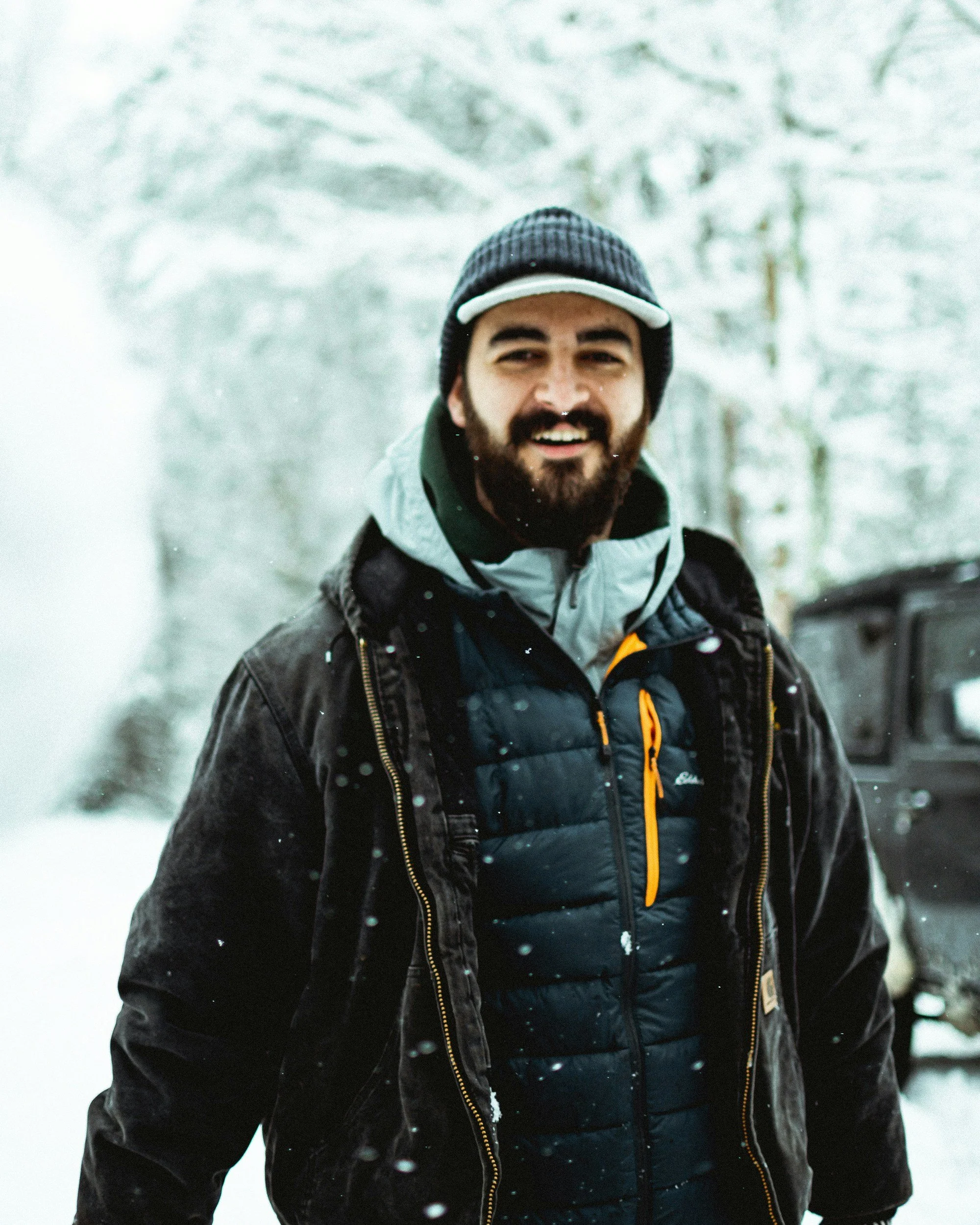 A man with a beard and wearing winter outdoor gear, including a beanie, jacket, and hoodie, standing in a snowy forest, smiling at the camera.