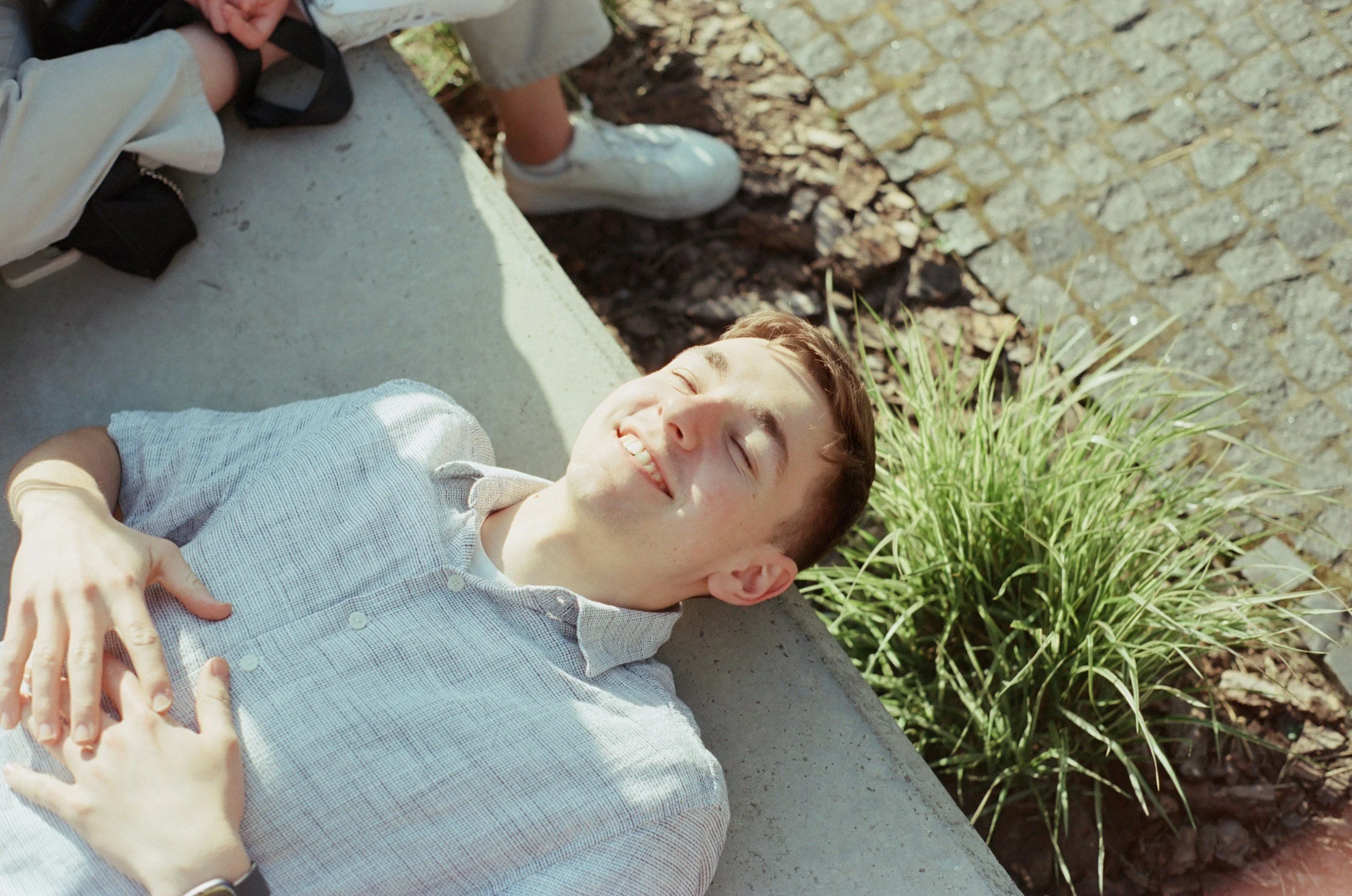 Young man lying on a concrete bench outdoors, smiling with eyes closed, hands resting on his stomach, wearing a light-colored, checkered button-up shirt, surrounded by grass and cobblestone pavement.
