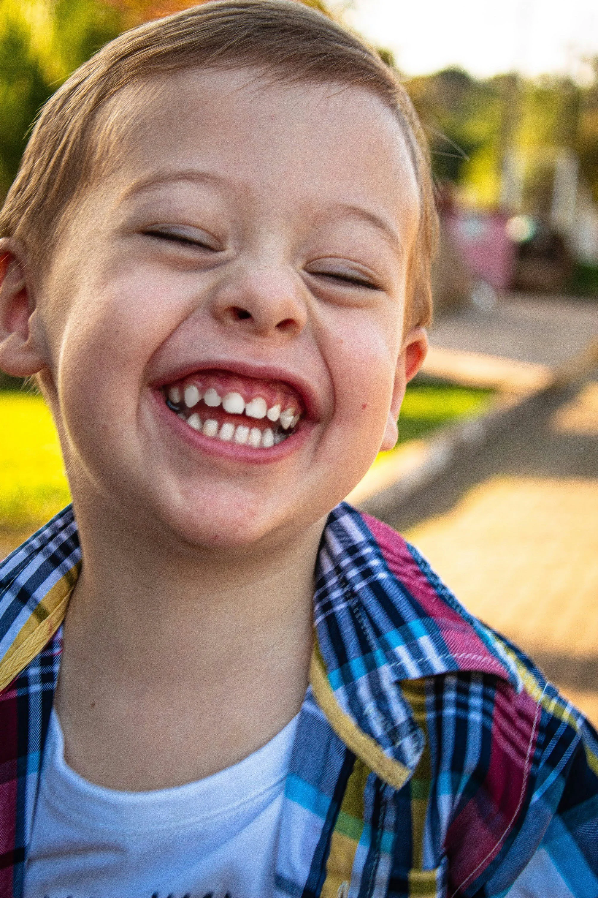 A young boy smiling brightly outdoors, showing missing teeth, wearing a colorful plaid shirt and a white undershirt.