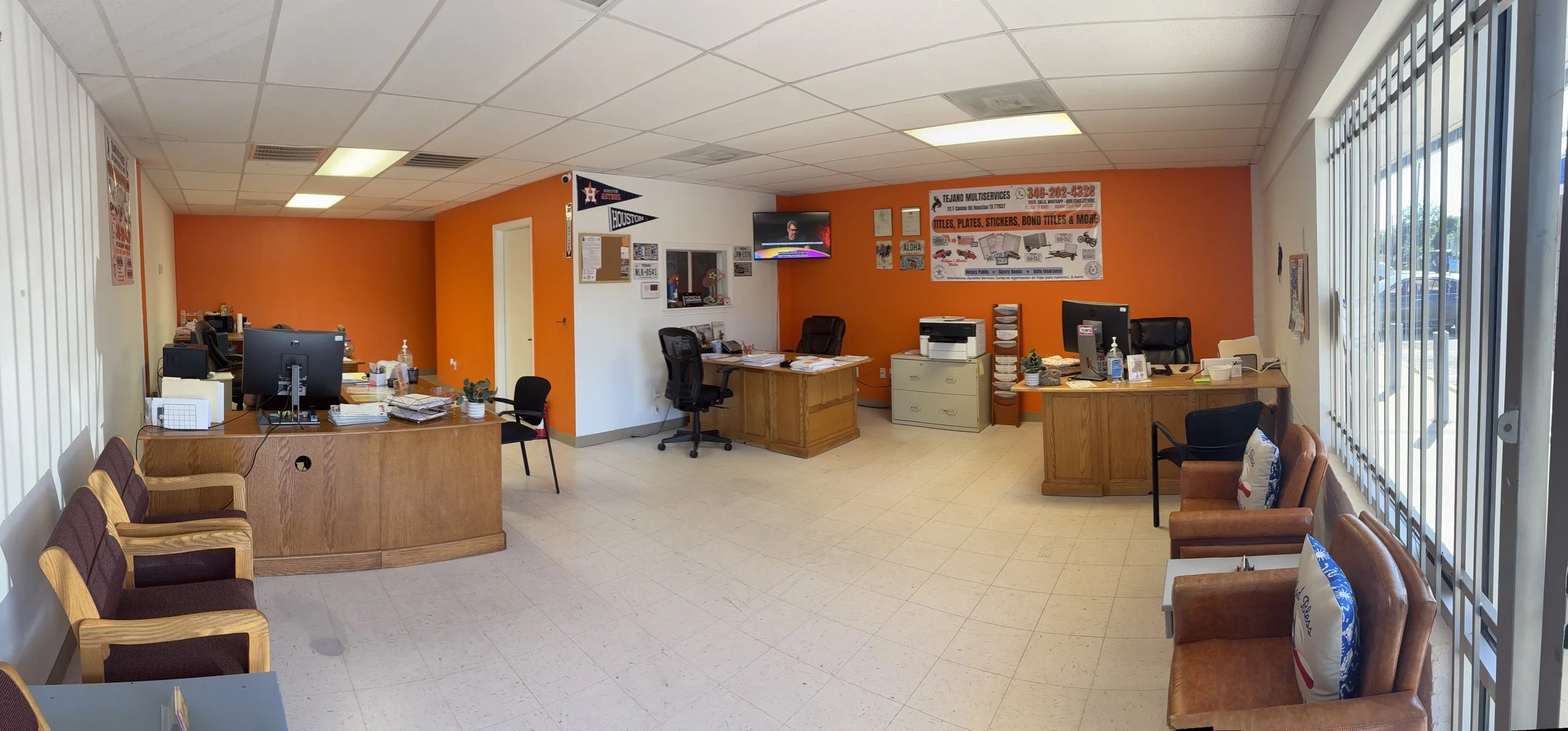 Office space with orange accent wall, two desks with computers, chairs, and office supplies, waiting area with chairs and pillows, large window with vertical blinds, TV mounted on wall, and various posters and signs.