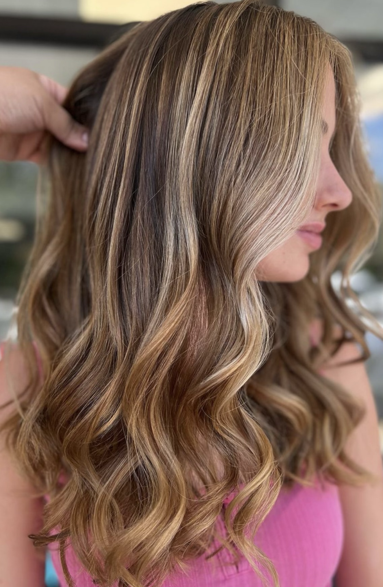 Close-up of a person with wavy, blonde balayage hair style, wearing a pink top.
