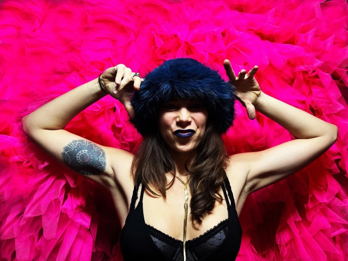 Dominicka, lying on a bright pink tutu, wearing a blue furry hat and dark lipstick, showing a tattoo on her arm