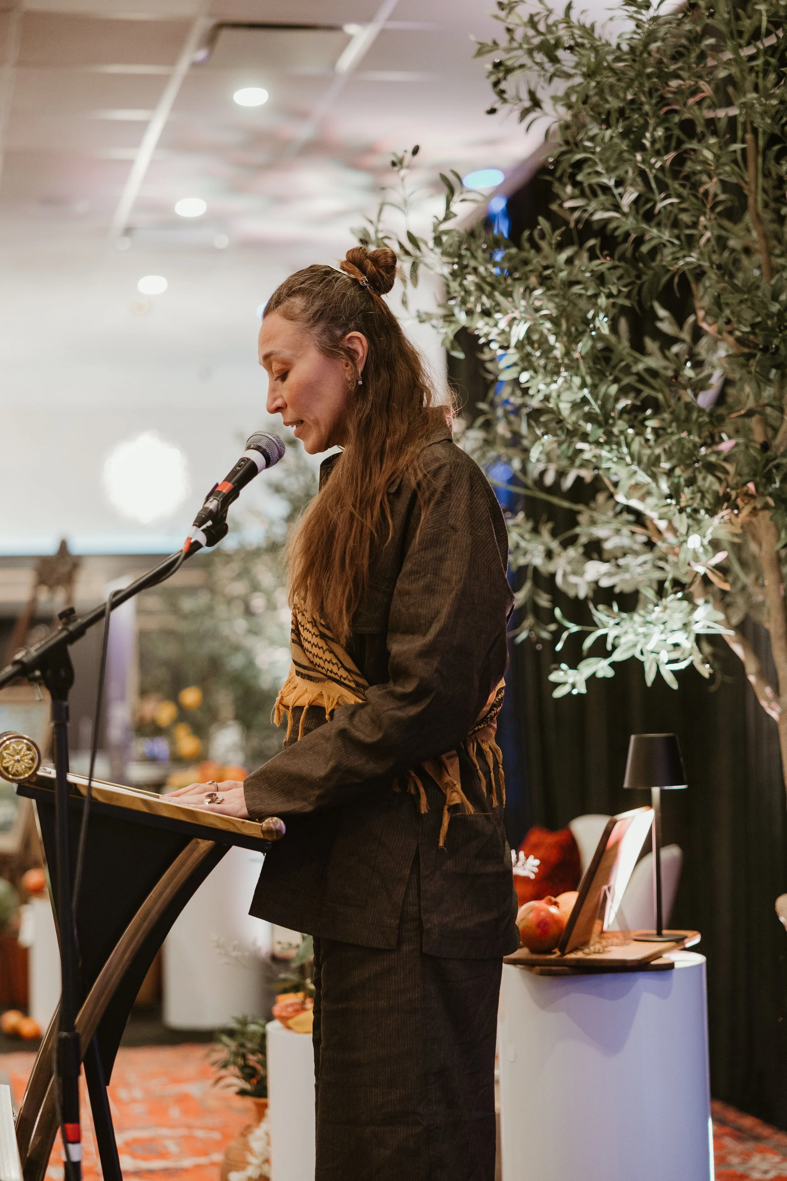 A woman with long brown hair tied in a top knot speaking into a microphone at a podium, wearing a dark jacket with a colorful woven scarf. Behind her is a leafy plant and a decorated display with fruits and a framed photo on a small table, in a well-lit indoor setting.