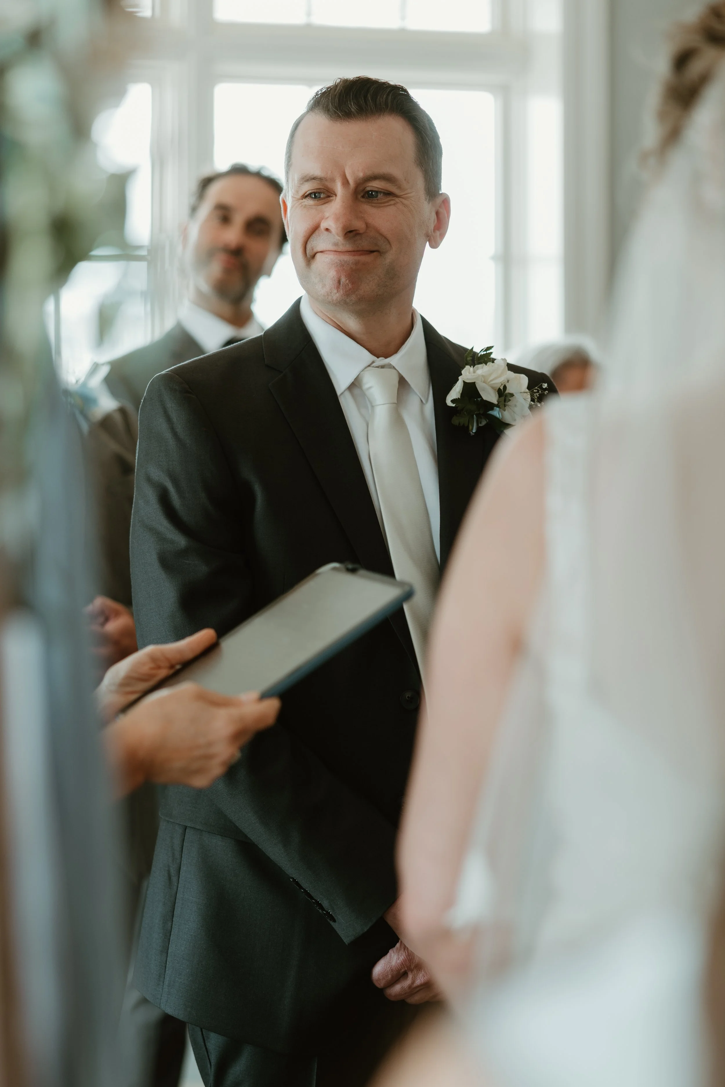 A groom in a black suit with a white tie and boutonniere looks at a bride during a wedding ceremony, with a man in a suit smiling in the background at the Lakehouse in Pickering, Ontario.