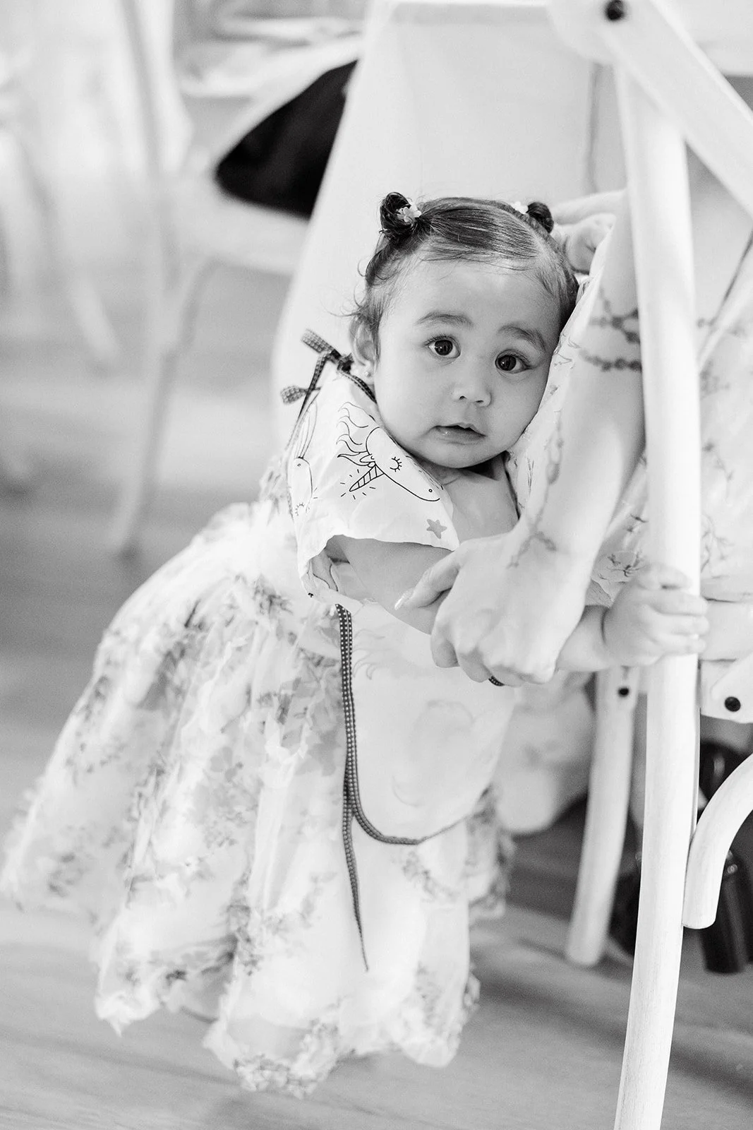 A young girl in a dress with a unicorn print is standing and holding onto a chair, looking at the camera at a baby shower in Mississauga, Ontario.