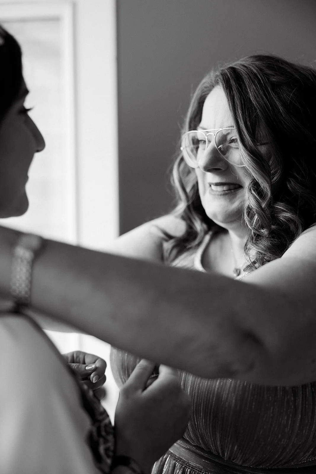 A woman with glasses and curly hair smiling as she adjusts another woman's shirt, who is out of frame. Black and white photo. Getting ready for a wedding in Oakville, Ontario
