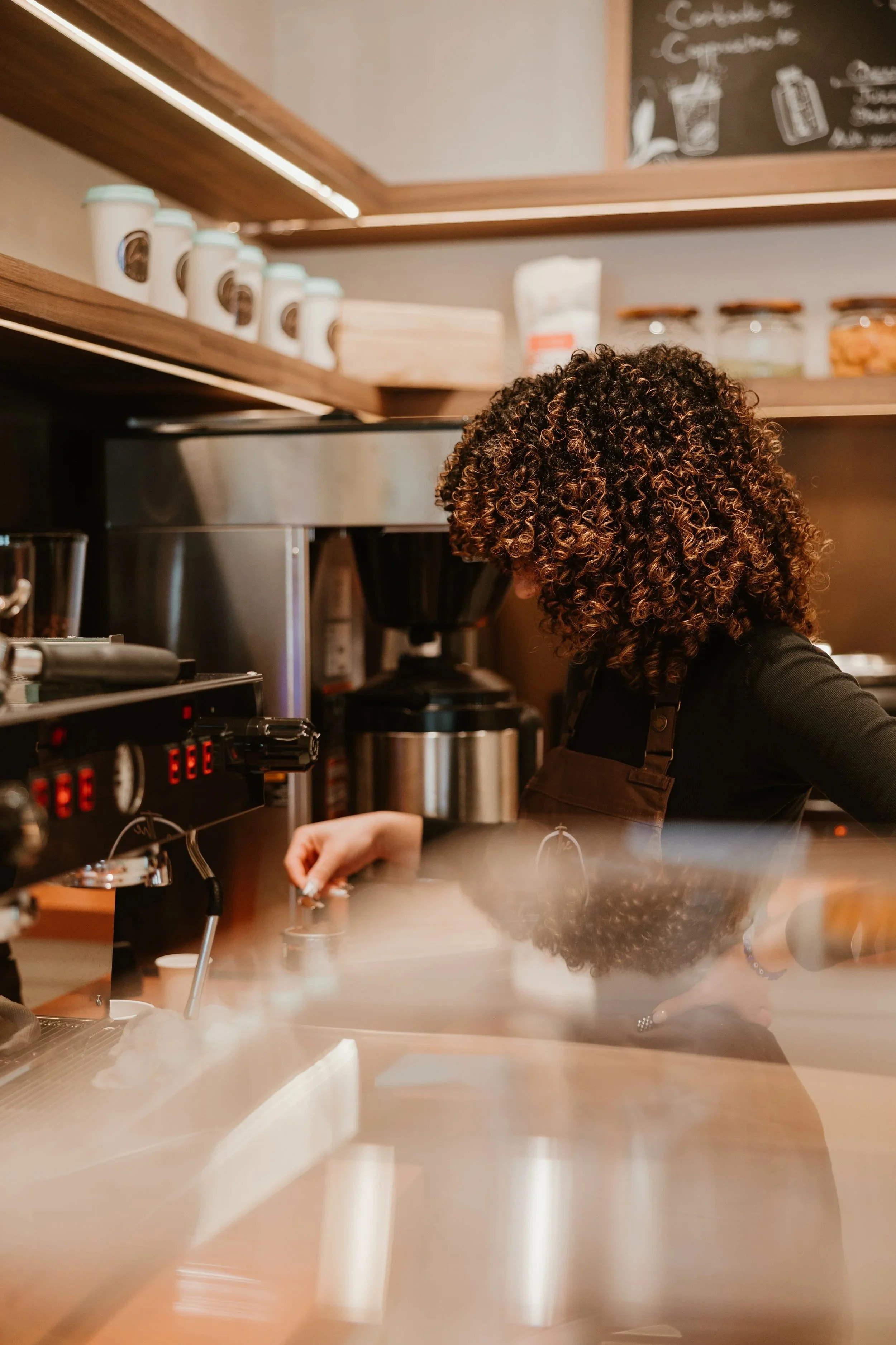 A woman with curly hair wearing a black shirt and apron working behind a coffee bar with espresso machines at Local by Masrawy in Mississauga, Ontario.