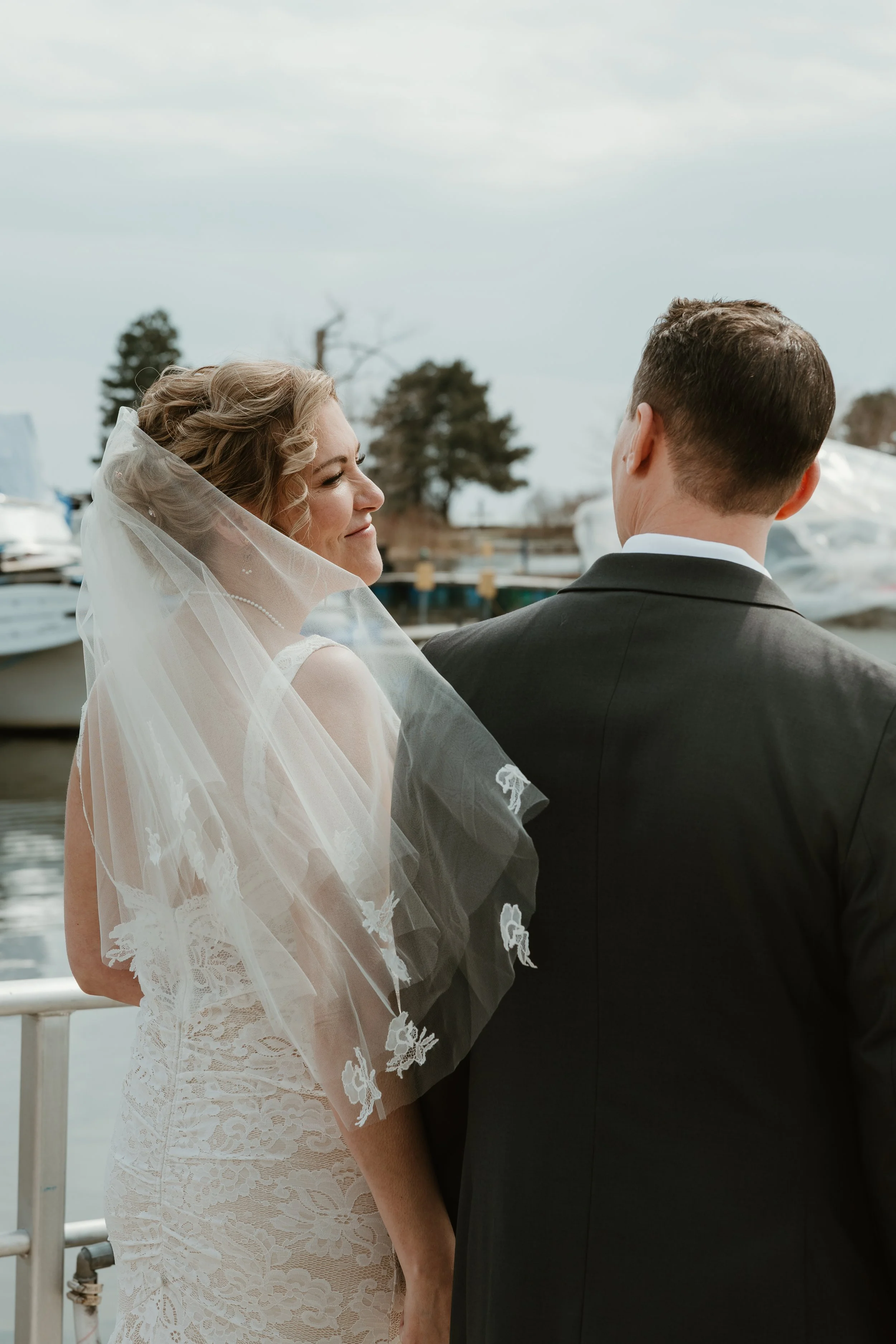 A bride and groom standing close together outdoors near a body of water, with trees and boats in the background, during daytime. From the Lakehouse in Pickering, Ontario.