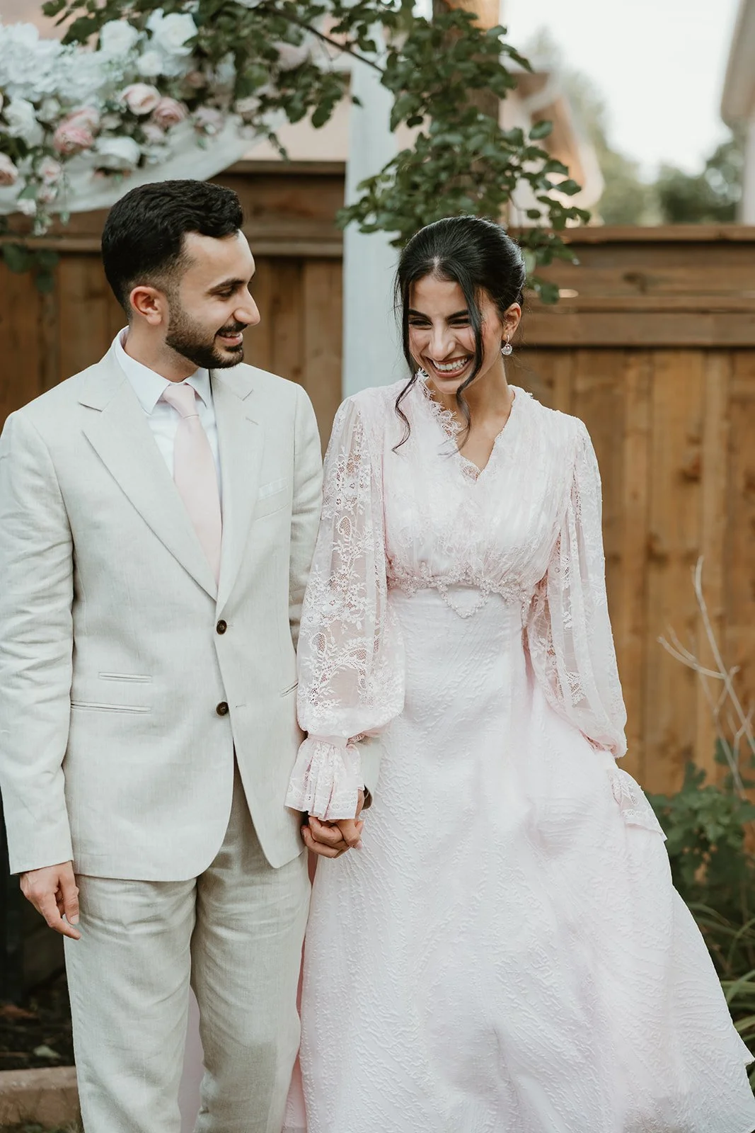 A bride and groom holding hands and smiling at each other outdoors, with a wooden fence and decorative flowers in the background.
